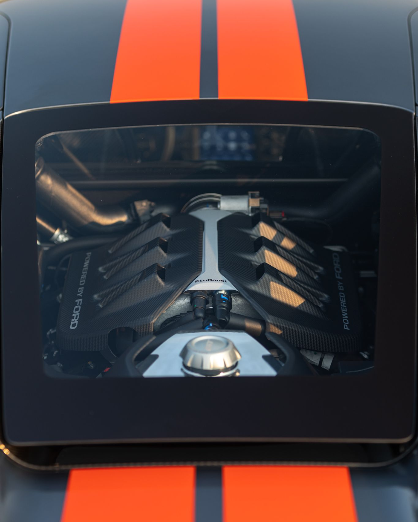 Black sports car hood with orange racing stripes and a clear window showing the engine underneath