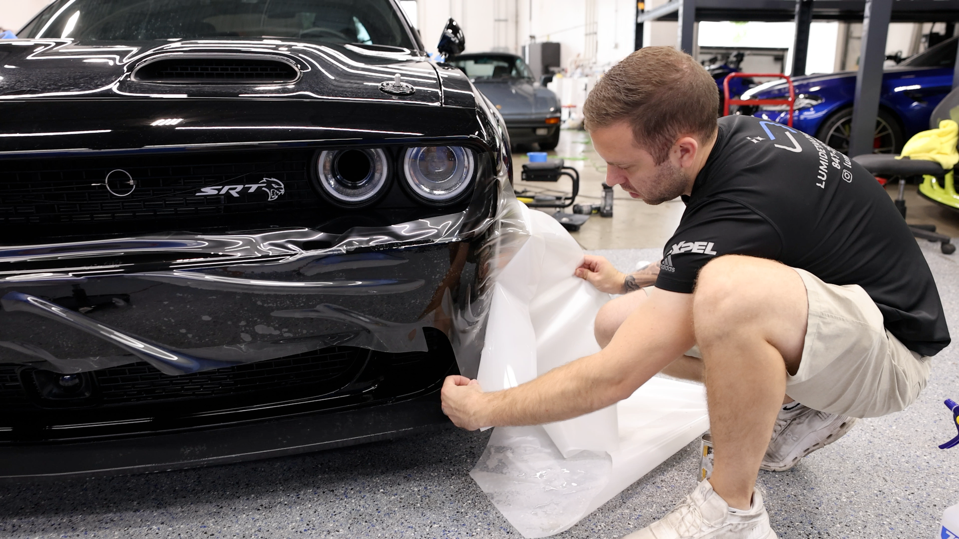 Man kneeling beside a black sports car, applying clear protective film in a workshop