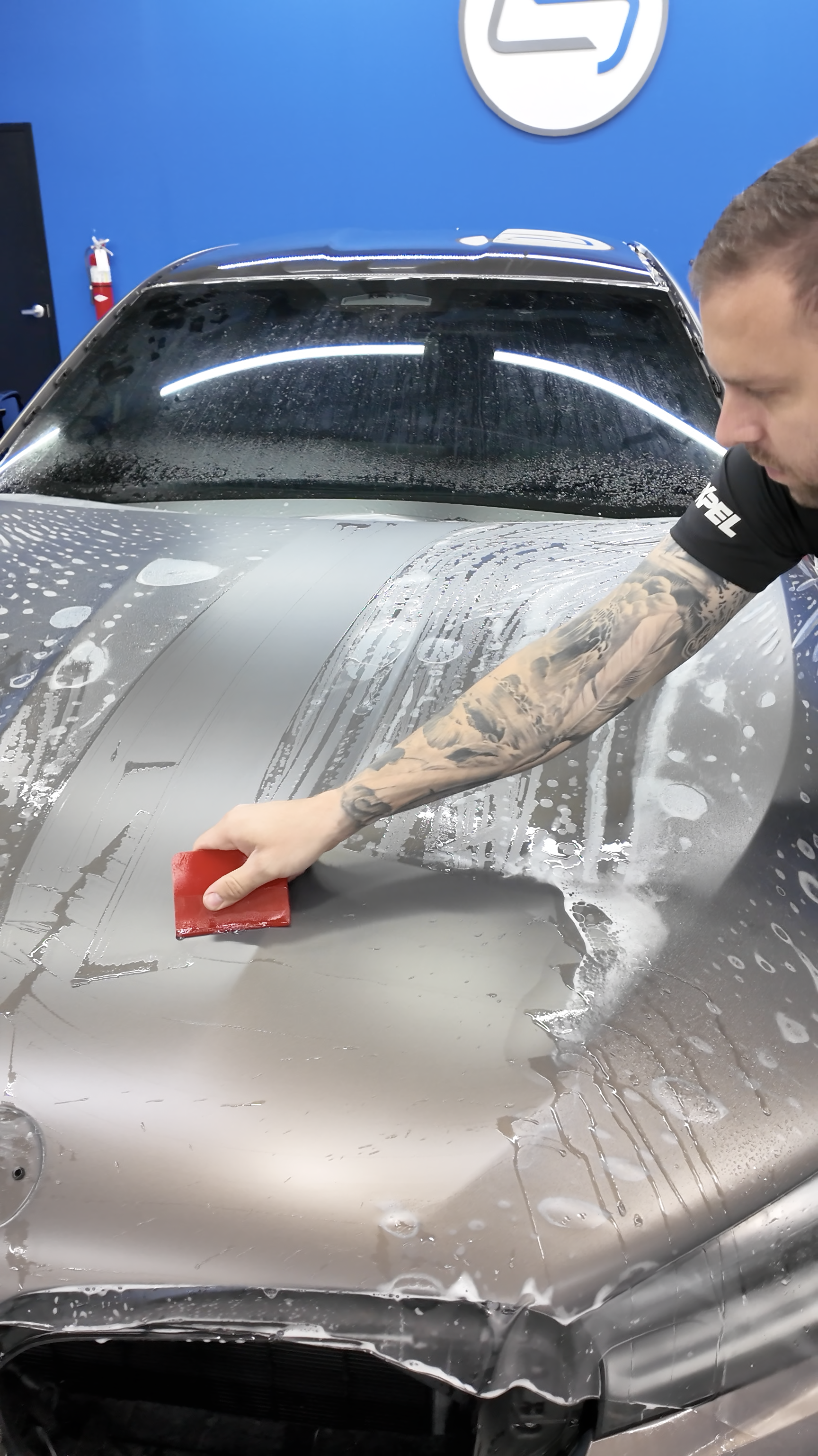 Person washing a gray car hood with a red sponge in a car wash, foam and water on the surface