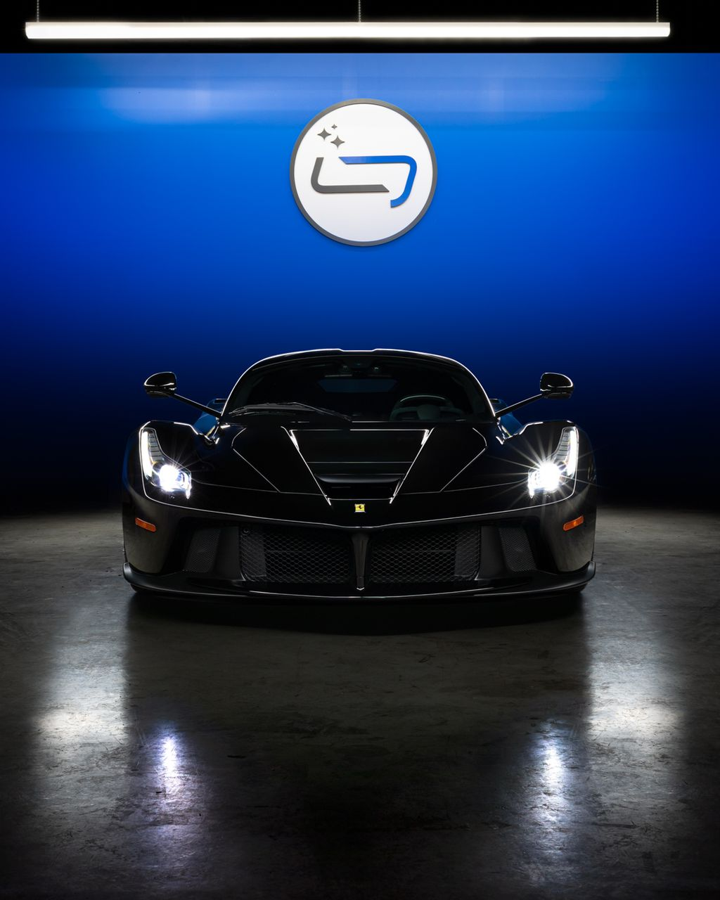 Black sports car in a showroom, facing forward under bright lights with a blue wall logo behind it