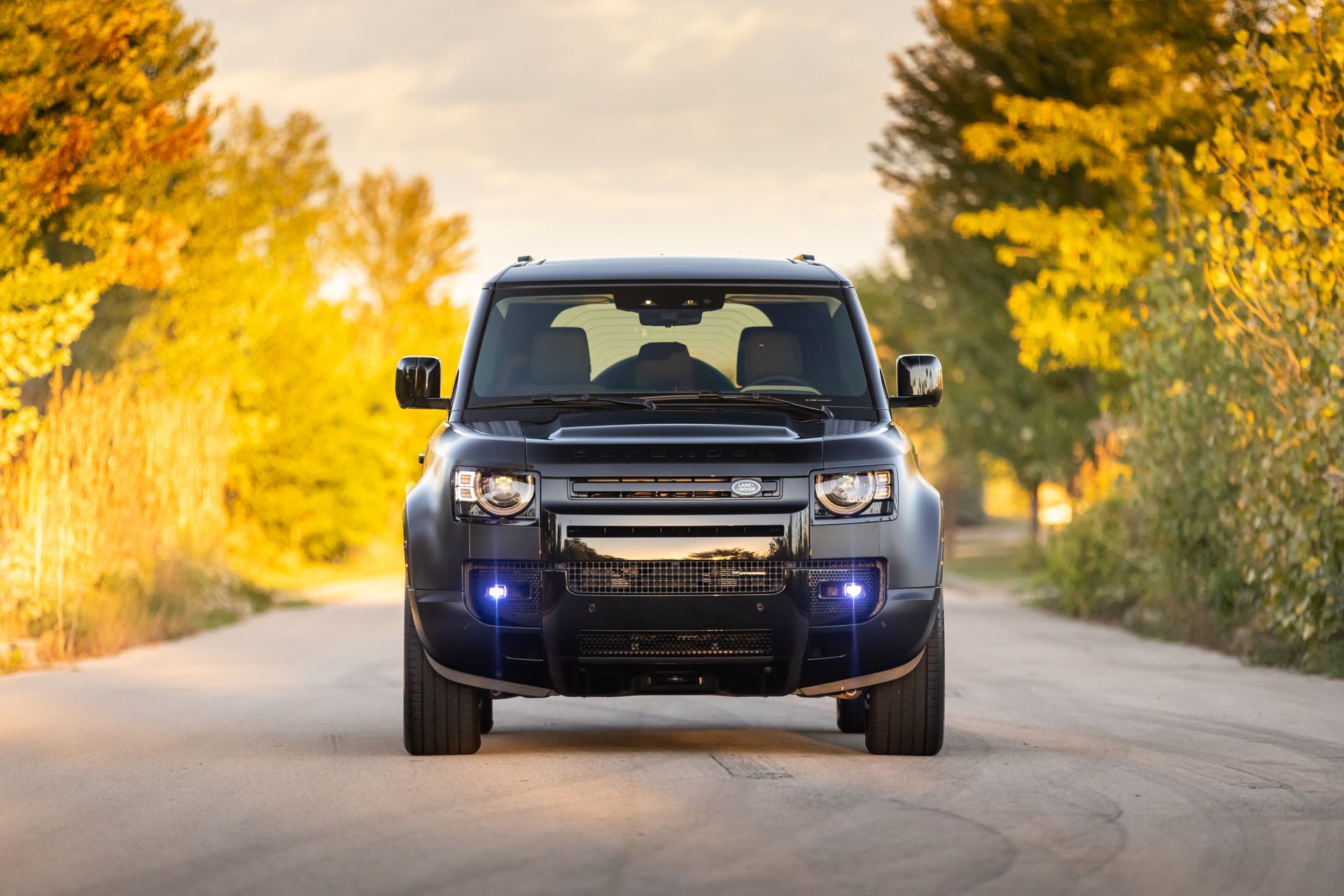 Black SUV driving on a tree-lined road in autumn, headlights on.