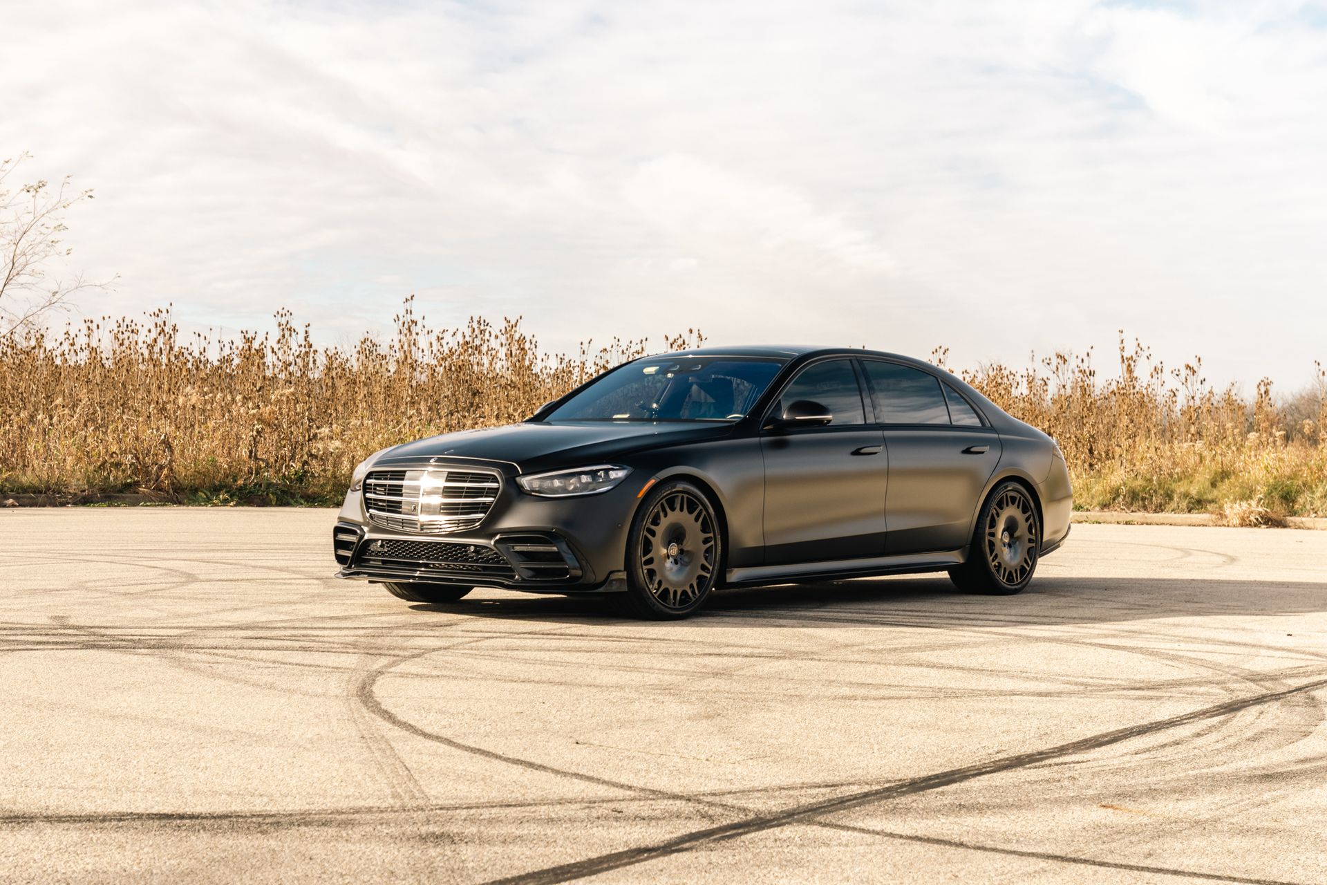 Black sedan parked on an empty paved lot beside dry reeds under a bright sky