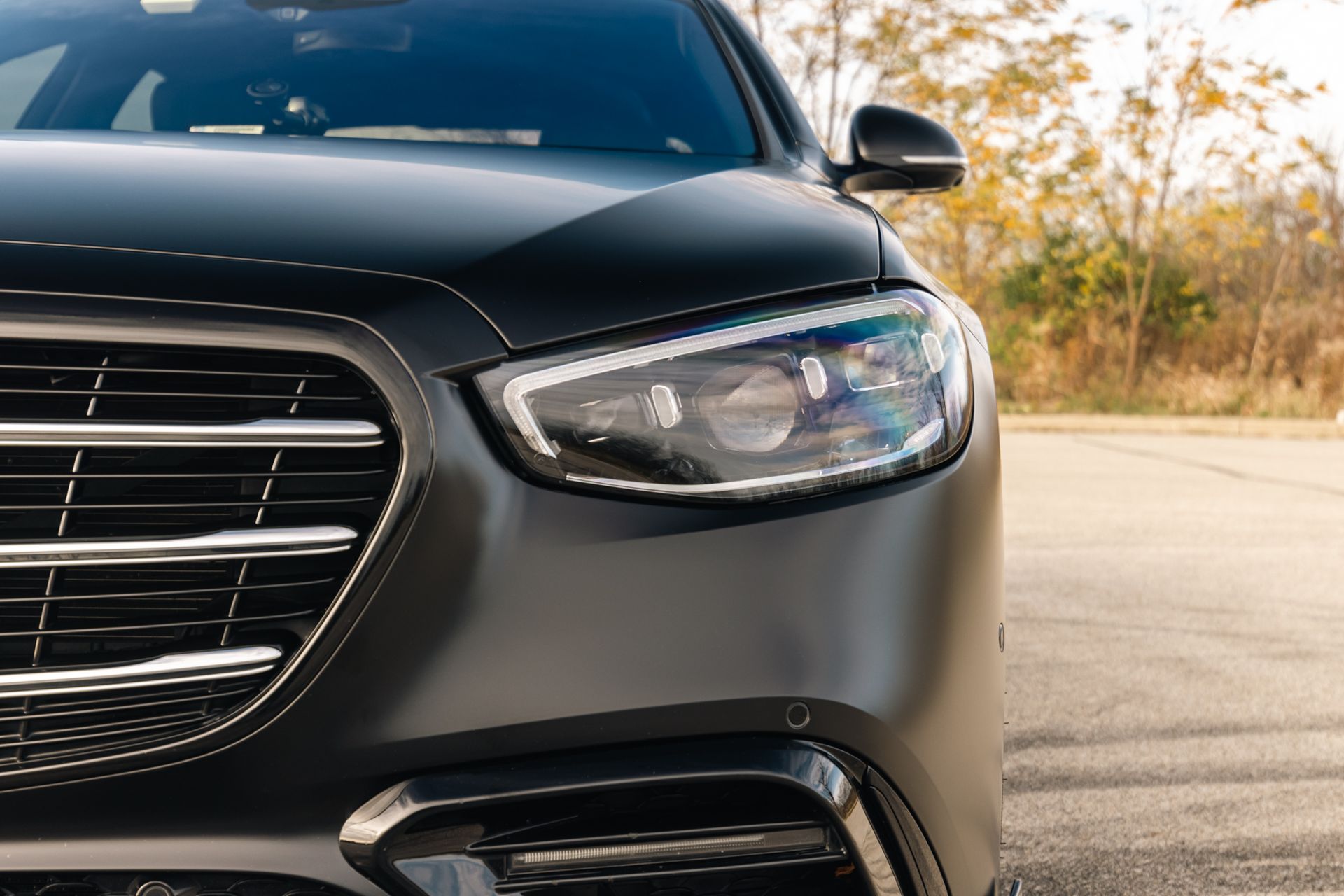 Close-up of a black sedan’s front headlight and grille on a road beside autumn trees