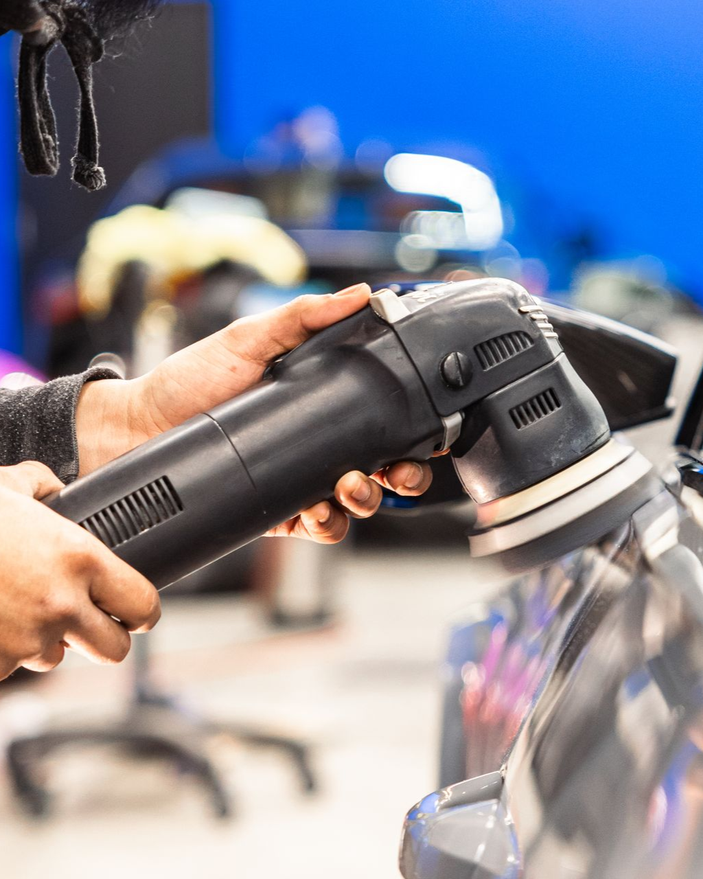 Person using a black rotary polisher on a car in a workshop, with a blue background.
