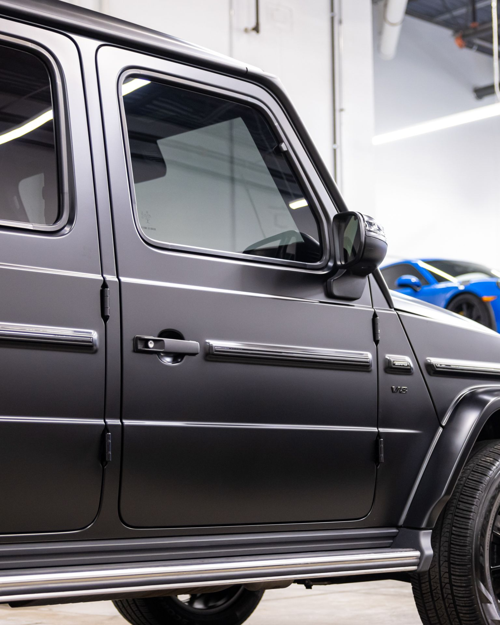 Side view of a black SUV with tinted windows parked indoors, with a blue sports car in the background