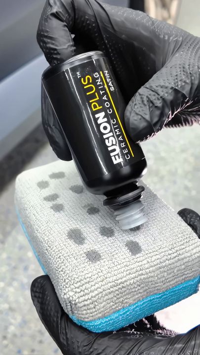 Gloved hands squeeze black Emulsion Plus bottle onto a gray microfiber cloth