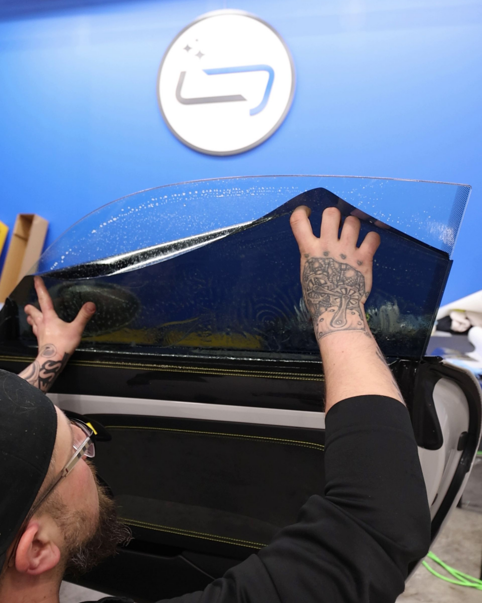 Person applying a blue protective film to a car hood in a workshop.