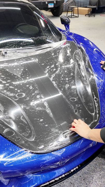 Person smoothing clear film on a blue sports car hood in a garage