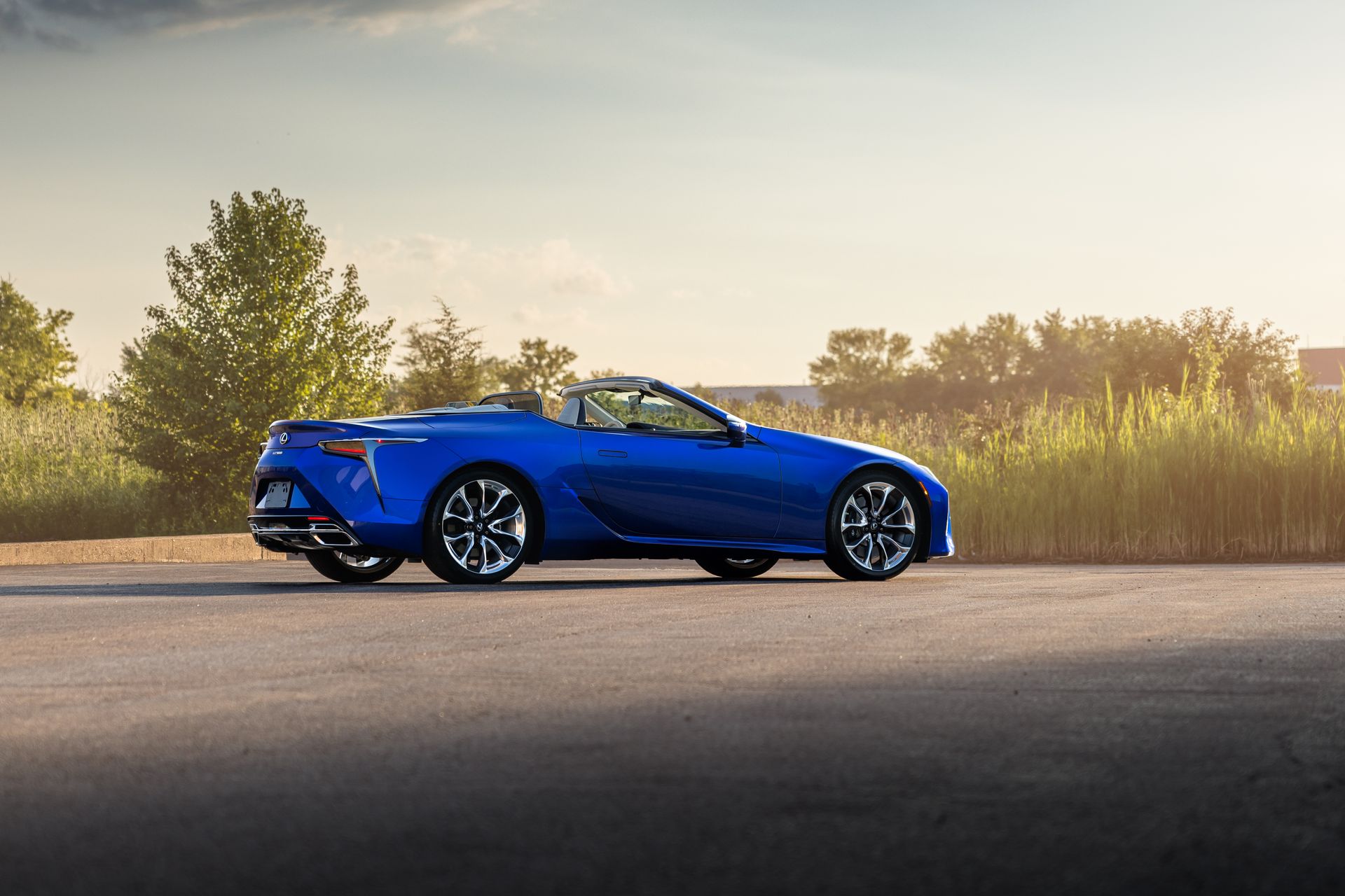 Blue sports car parked on a road beside tall grass at sunset