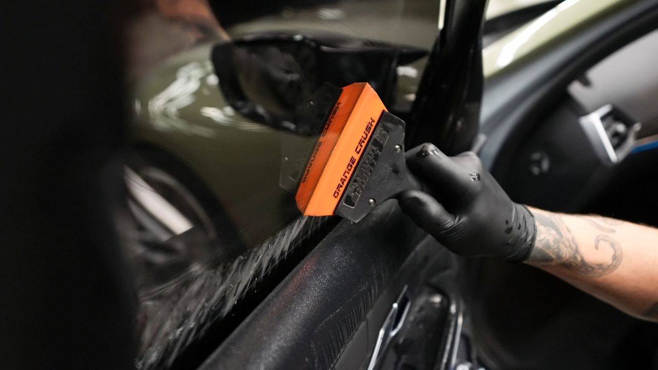 Person using an orange squeegee to apply film on a car window during tinting or wrapping