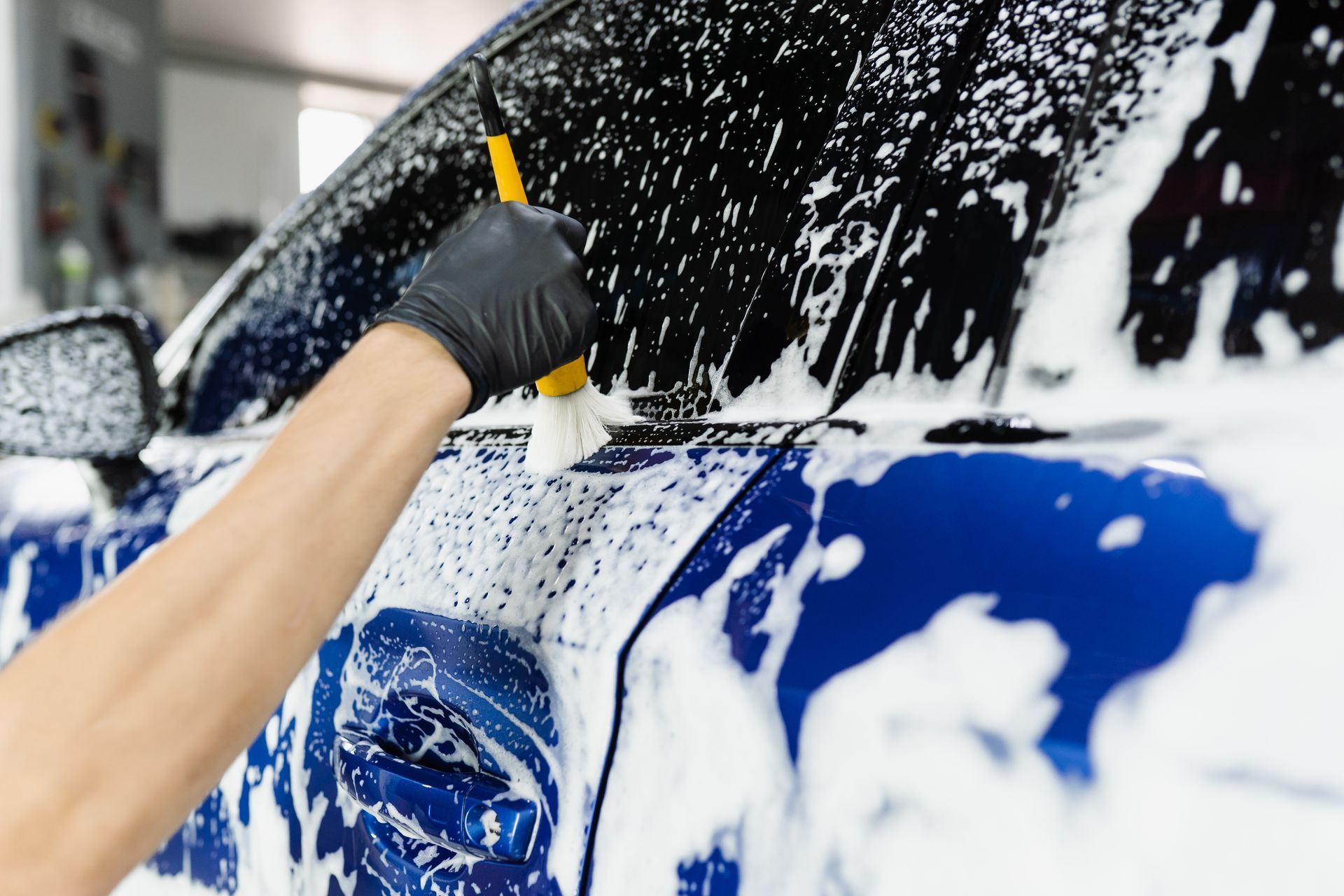 Gloved hand washing a blue car with soap foam in an auto detailing bay