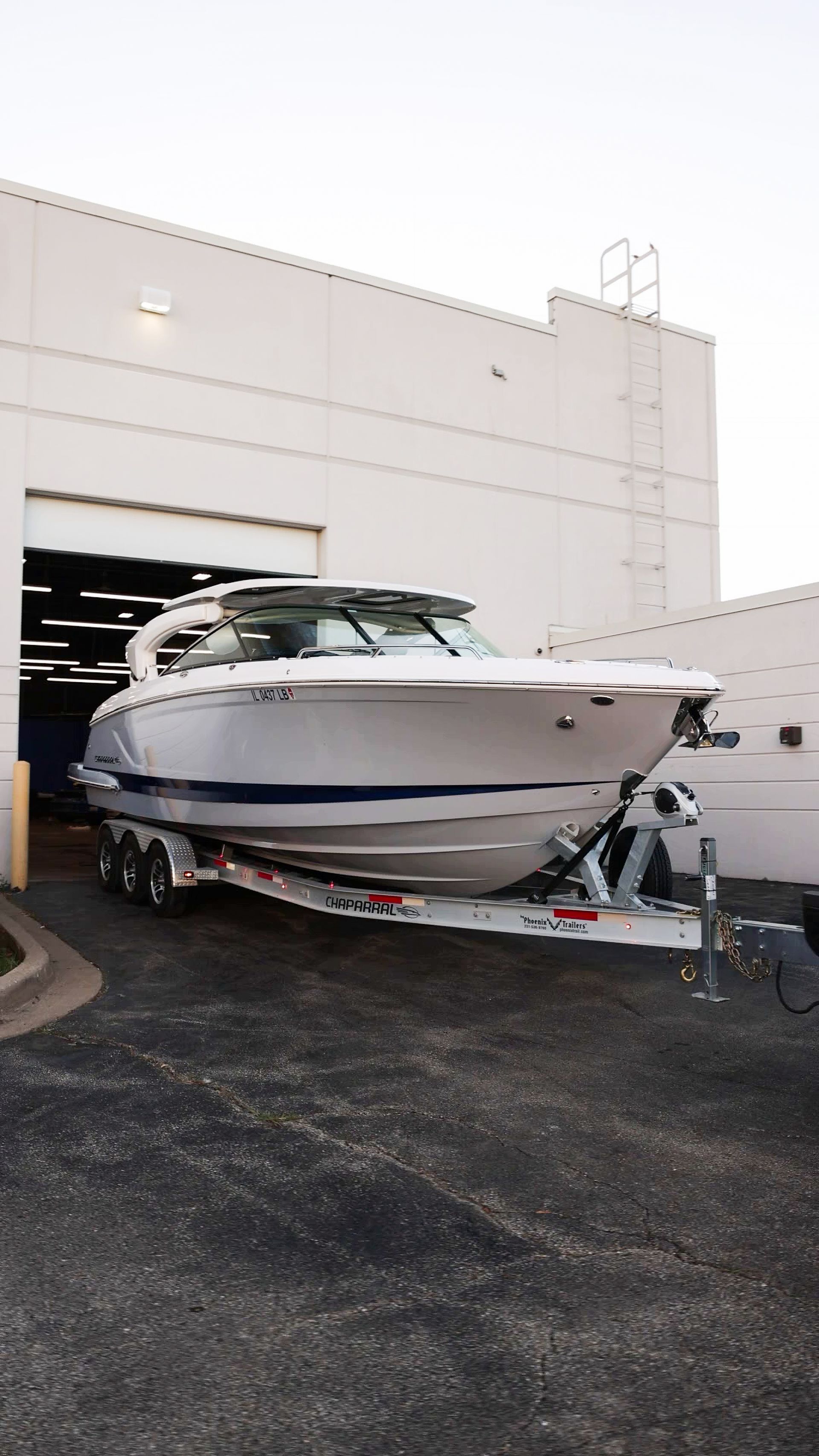 White motorboat on a trailer parked outside a large industrial building.