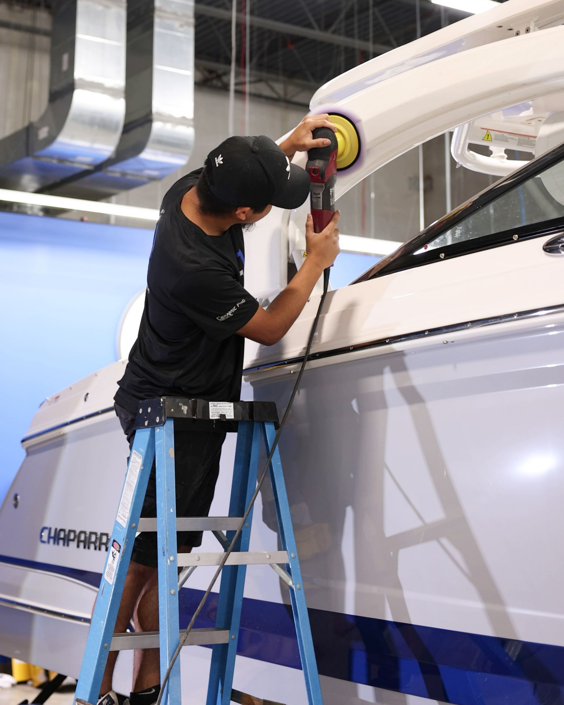 Worker sanding a white vehicle panel with a power sander while standing on a step ladder.