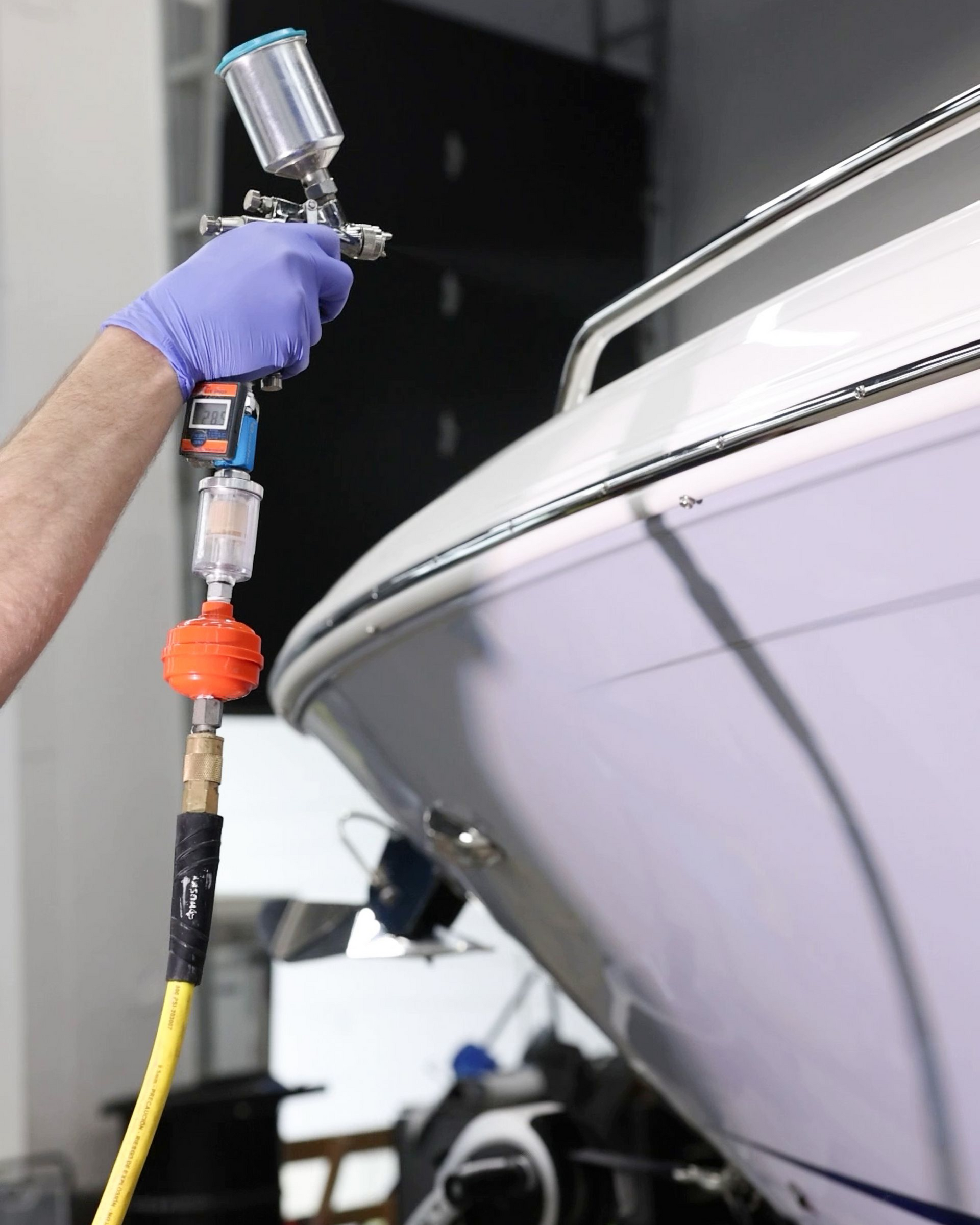 Gloved hand spraying a white boat hull with a spray gun on a workshop stand