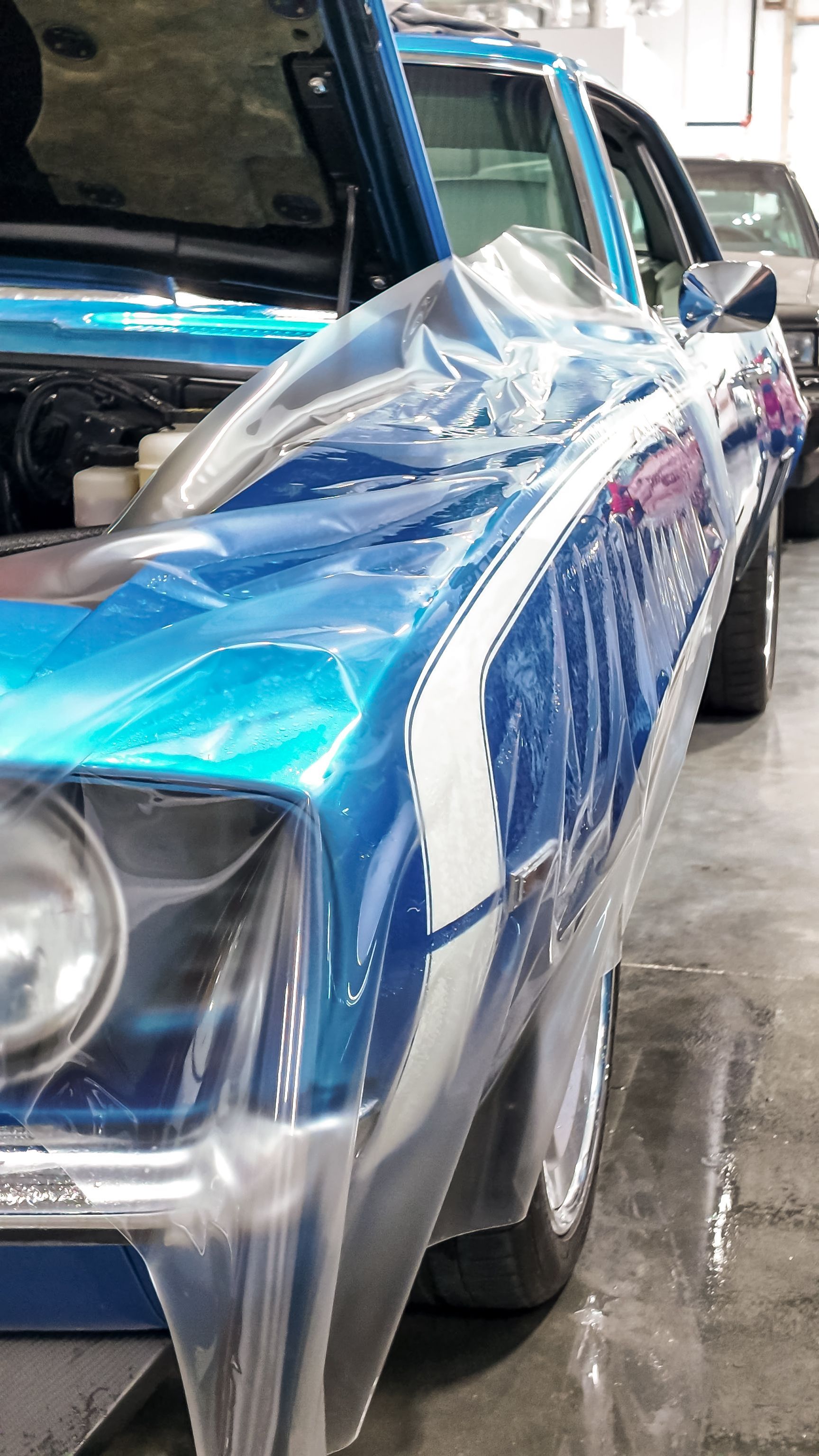 Blue vintage car wrapped in clear plastic in a garage, with a black SUV beside it