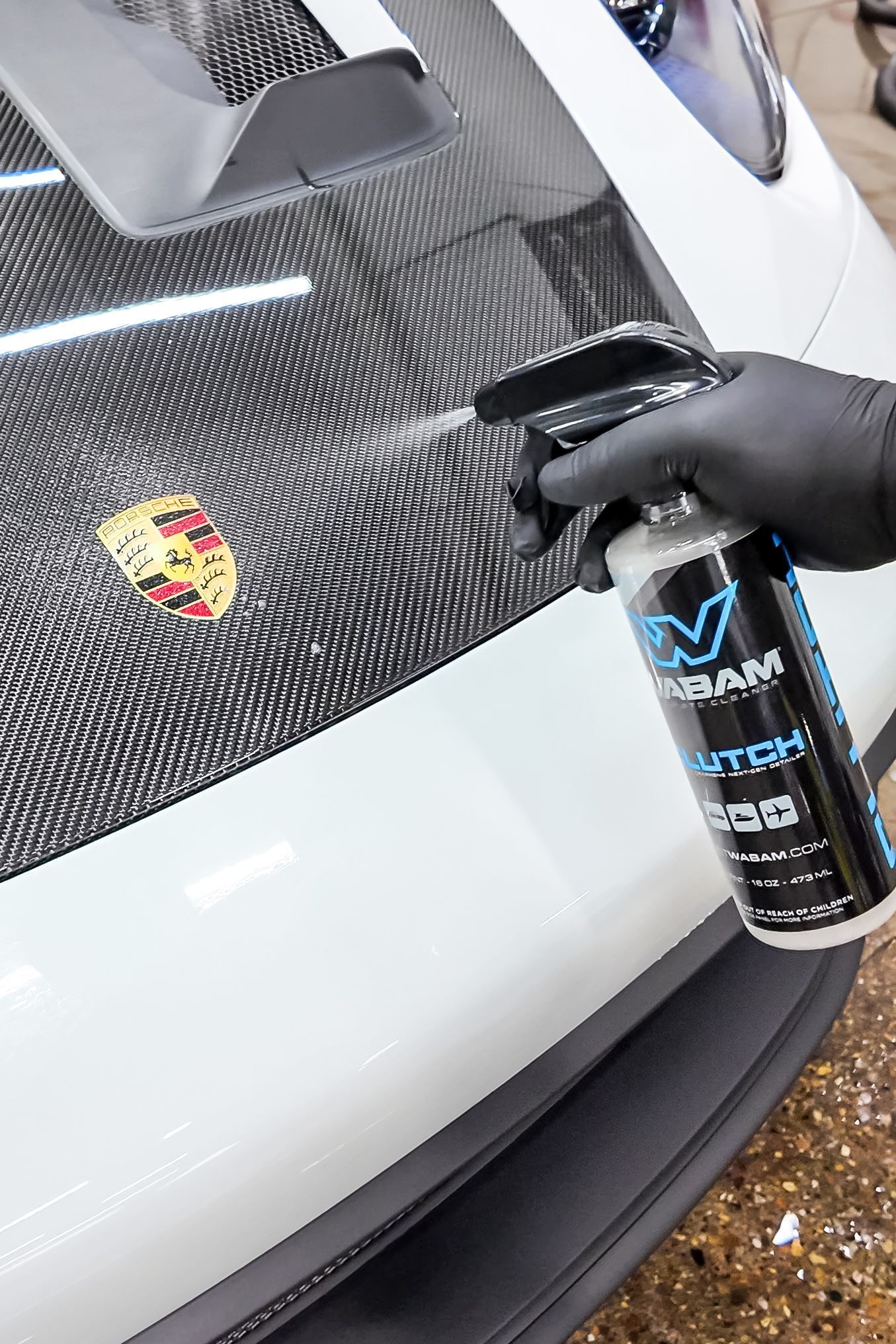 Gloved hand spraying a black carbon-fiber Porsche hood emblem with detailing spray