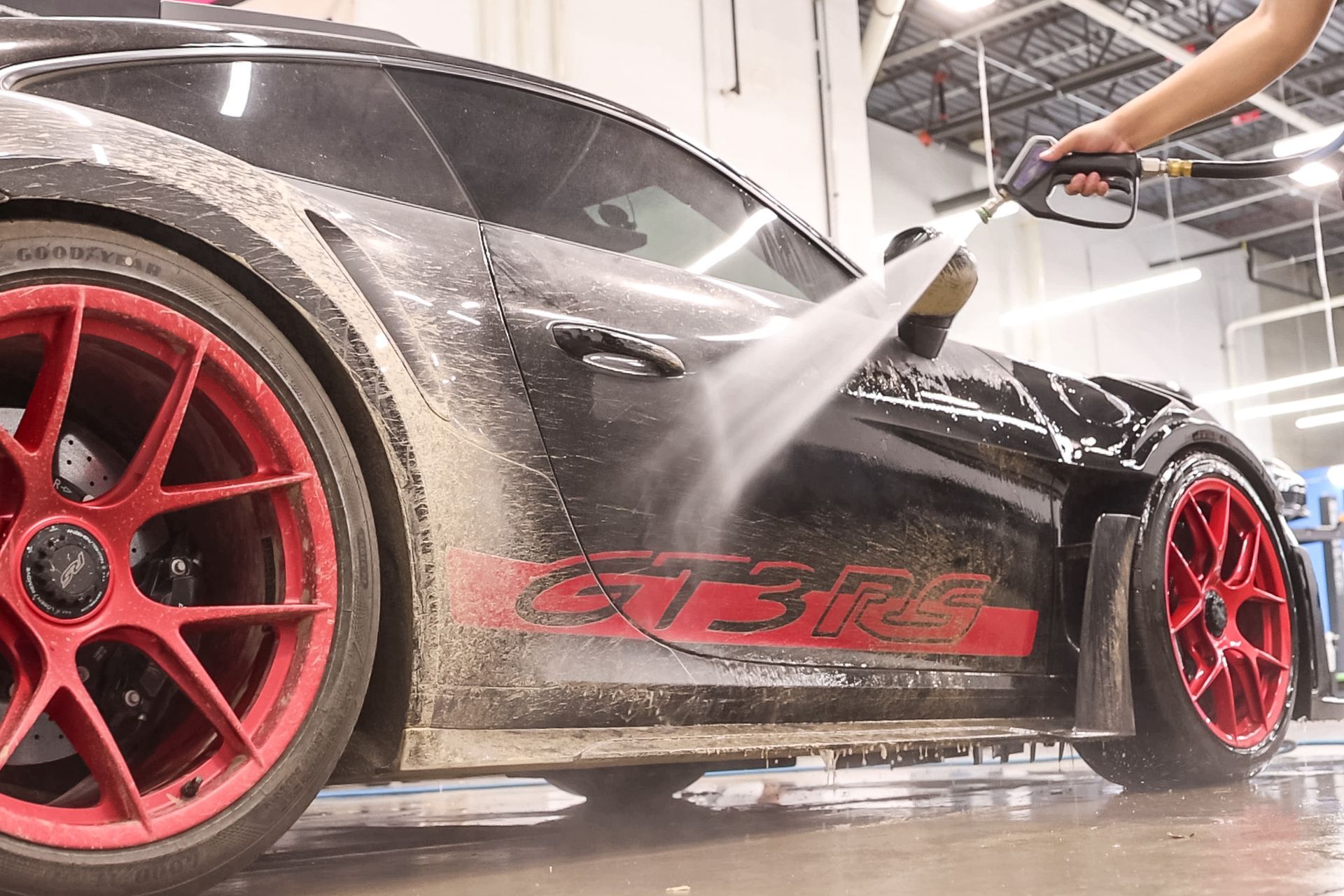 Red sports car being pressure washed in a garage, with large red rims and water spraying across the side.