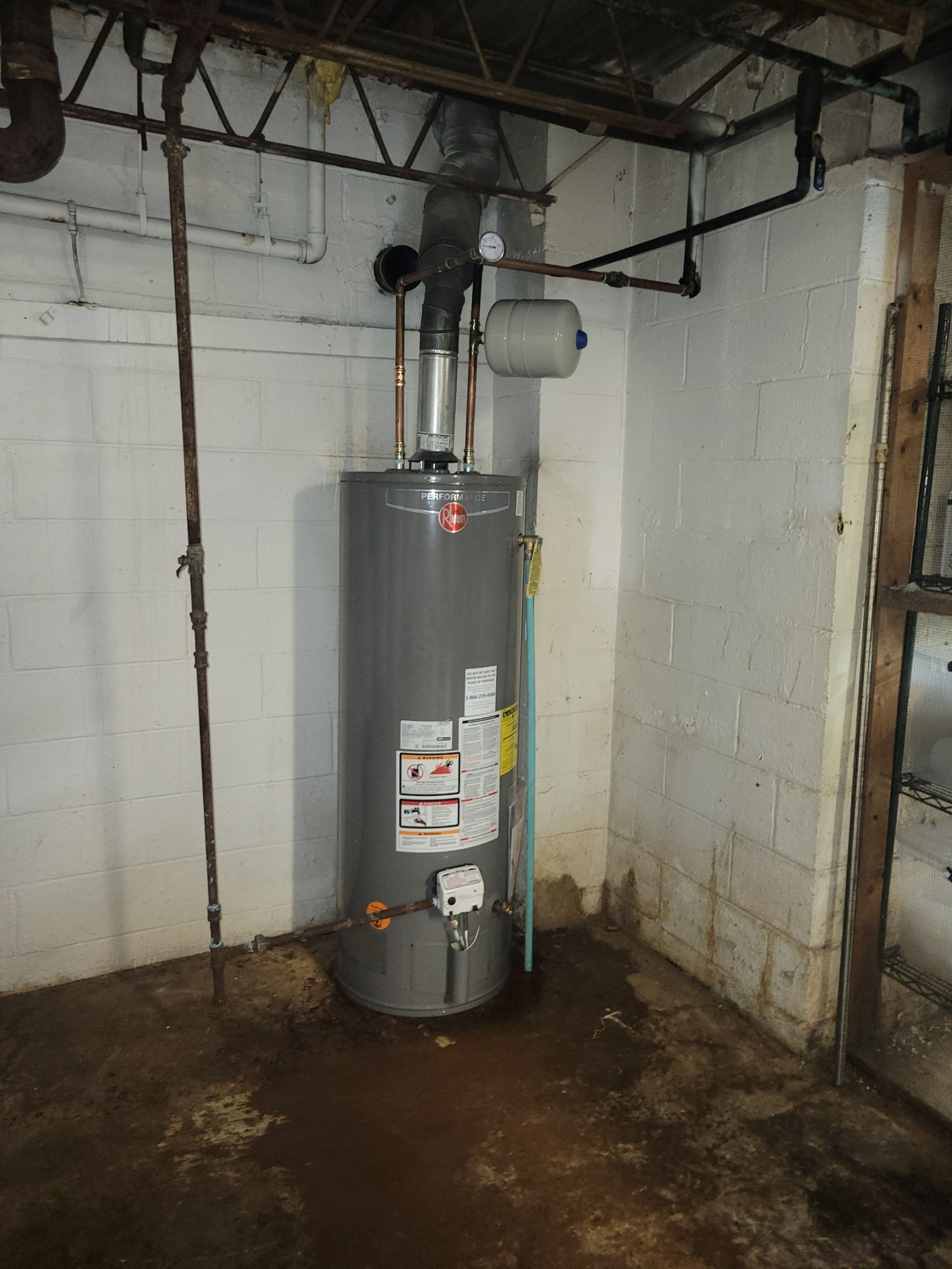 A gray water heater with a vent pipe stands in a corner against white cinder block walls on a damp concrete floor.