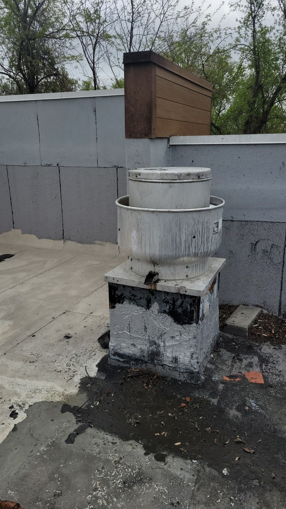 A rooftop exhaust fan sits on a painted masonry base atop a flat, grey roof, with a wooden structure in the background.
