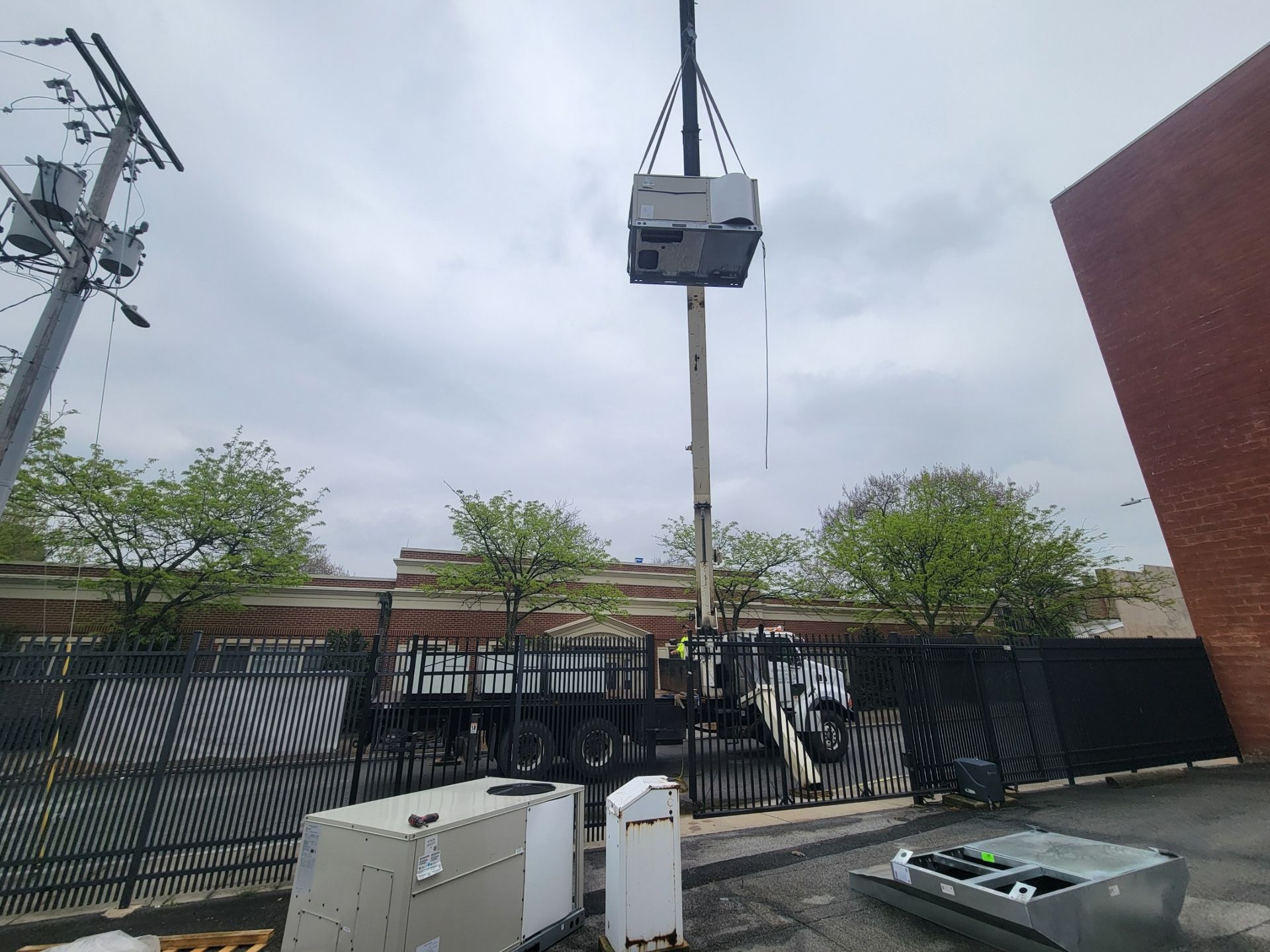 A crane lifts a large HVAC unit in an urban parking lot next to a brick building.