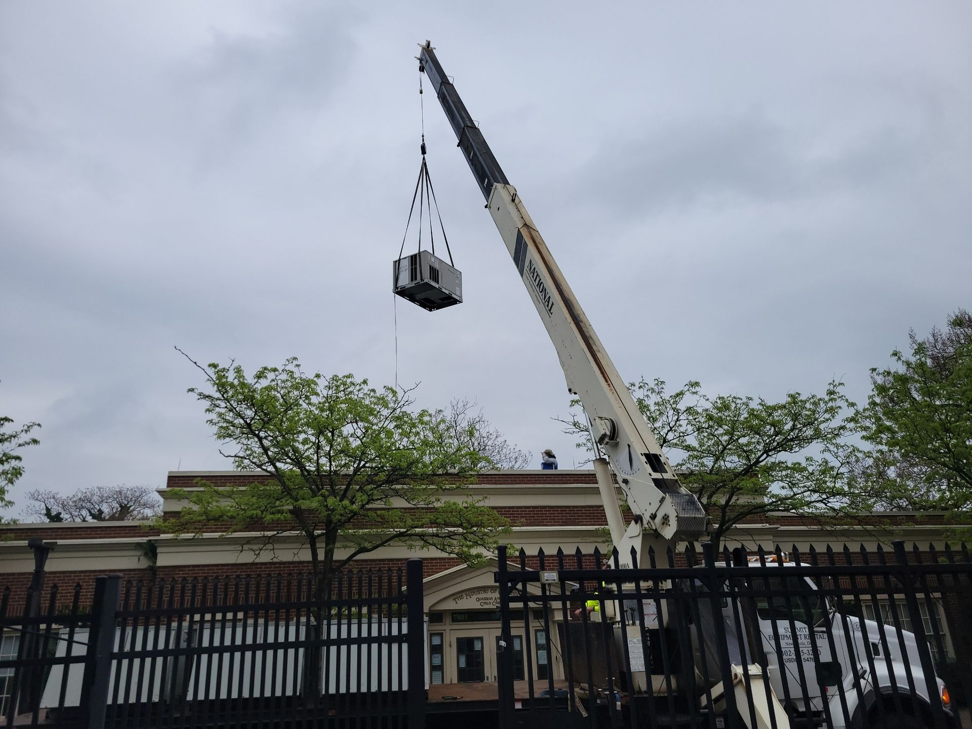 A crane lifts a large industrial HVAC unit above a brick building behind a metal fence under a cloudy sky.