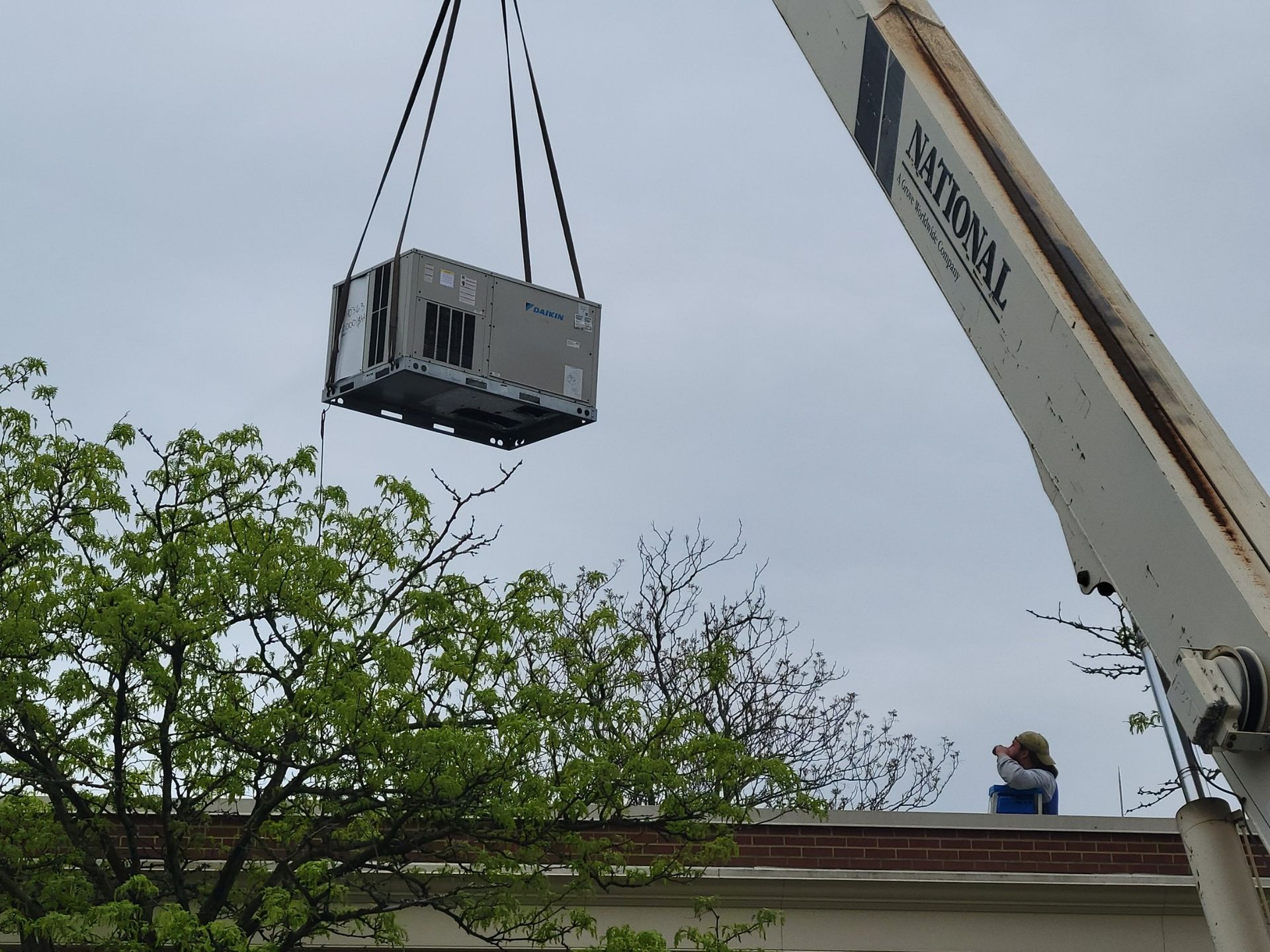 A crane lifts a large industrial HVAC unit through the air toward a building roof, with a worker standing nearby.