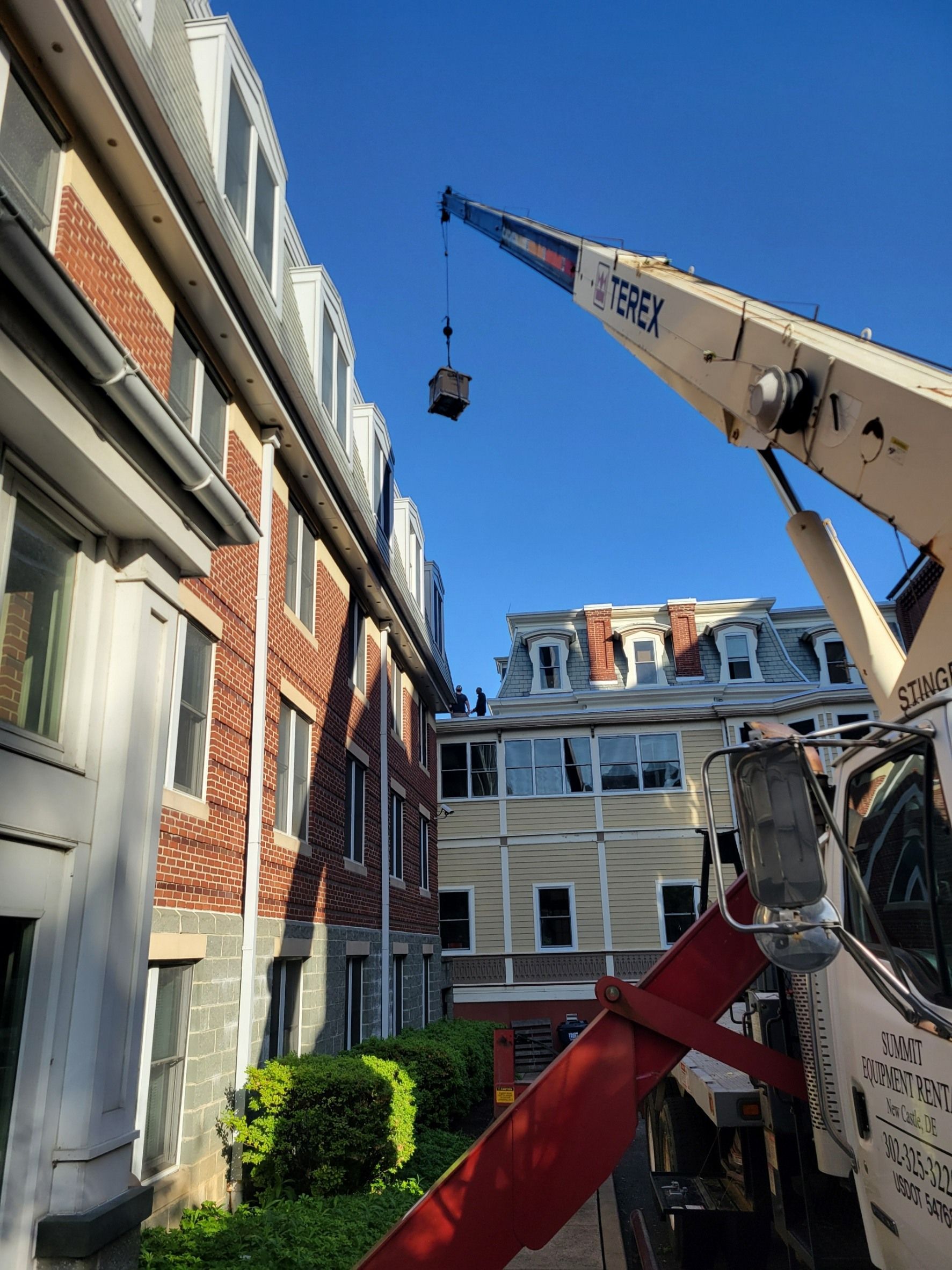 A crane lifts an object toward the roof of a brick apartment building under a clear blue sky.