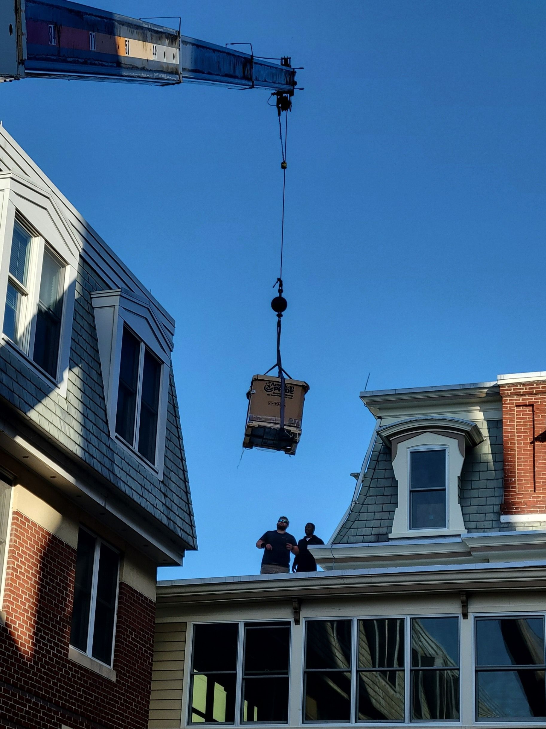 A crane lowers a package to workers on the flat roof of a building against a clear blue sky.