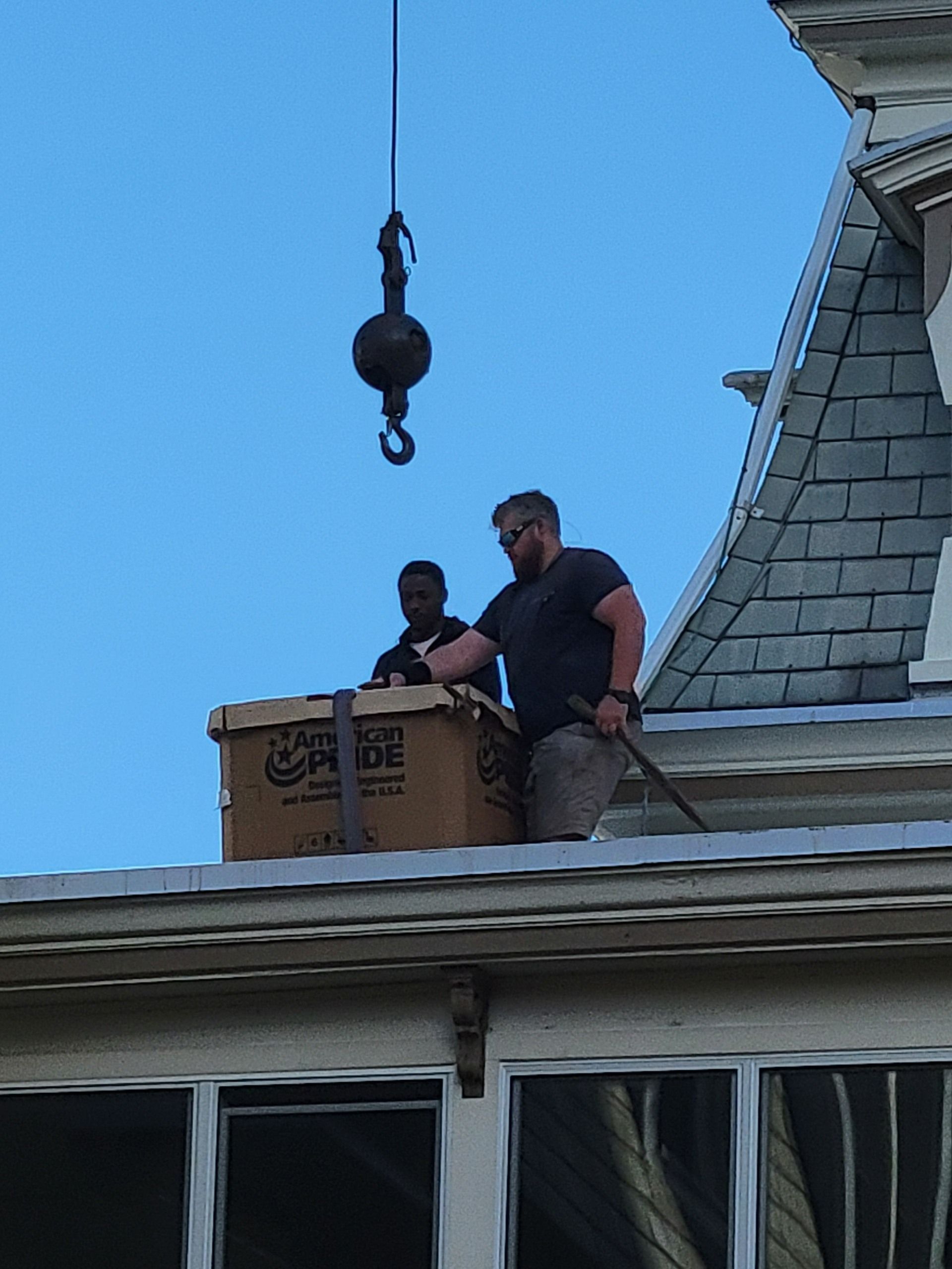 Two people on a roof guide a crated item suspended by a crane hook against a blue sky.