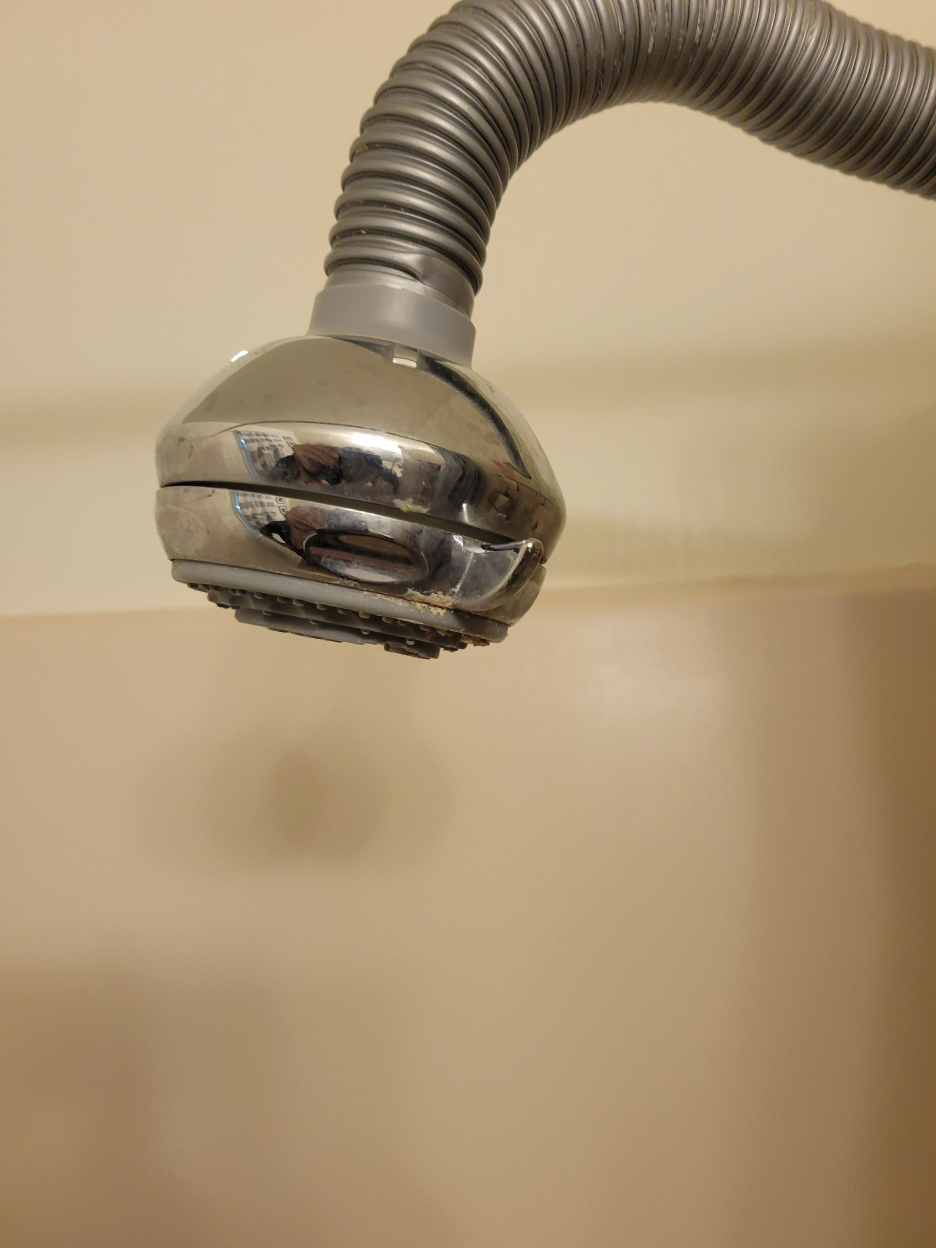 A close-up view of a metal shower head attached to a flexible, ribbed hose against a plain, beige-toned wall.