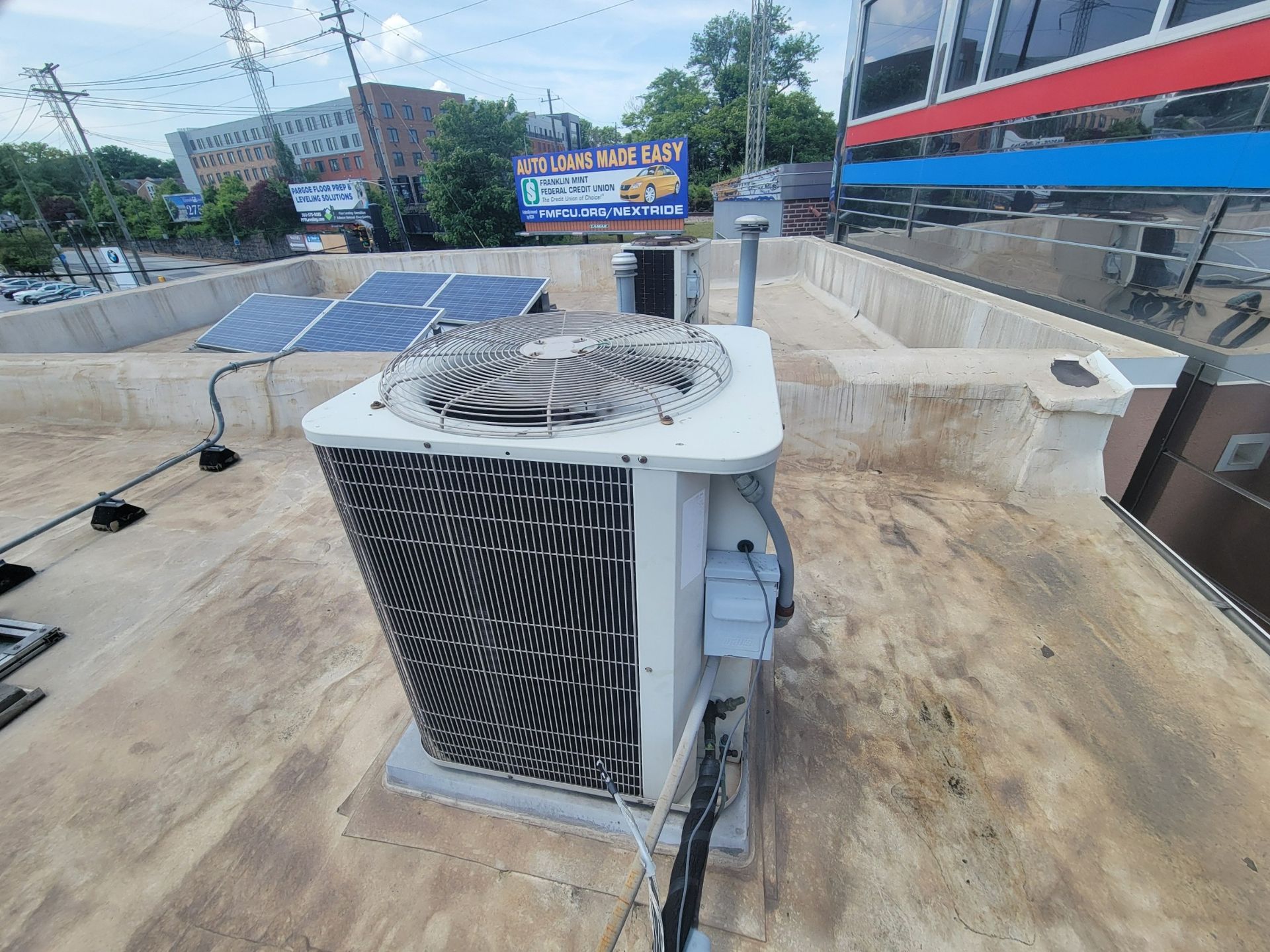 A white air conditioning condenser sits on a flat, tan rooftop, with solar panels and a distant billboard in the background.