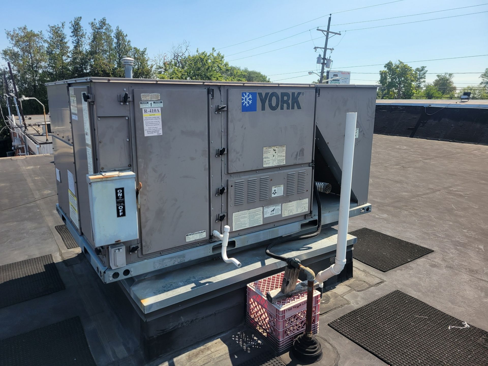 A gray York rooftop HVAC unit sits on a flat roof, with a small plastic crate and pipes located near the base.
