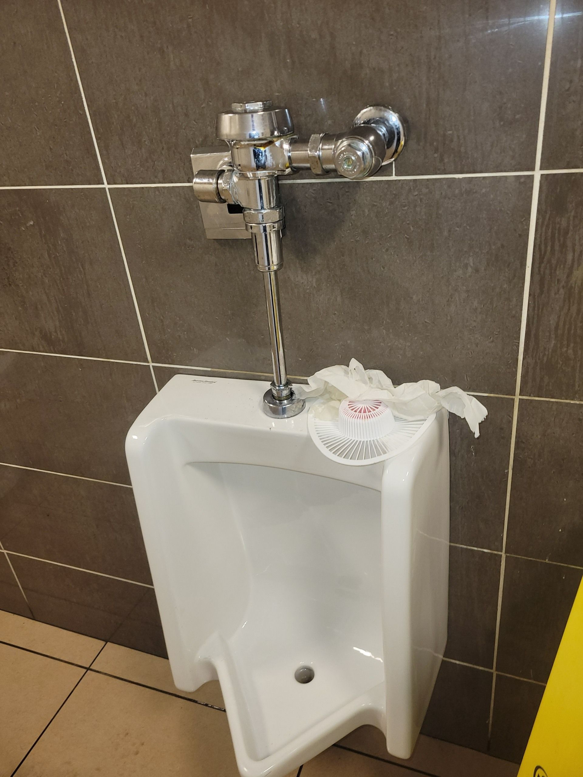 A wall-mounted white urinal with a paper towel draped over its top edge, set against a dark gray tiled wall.