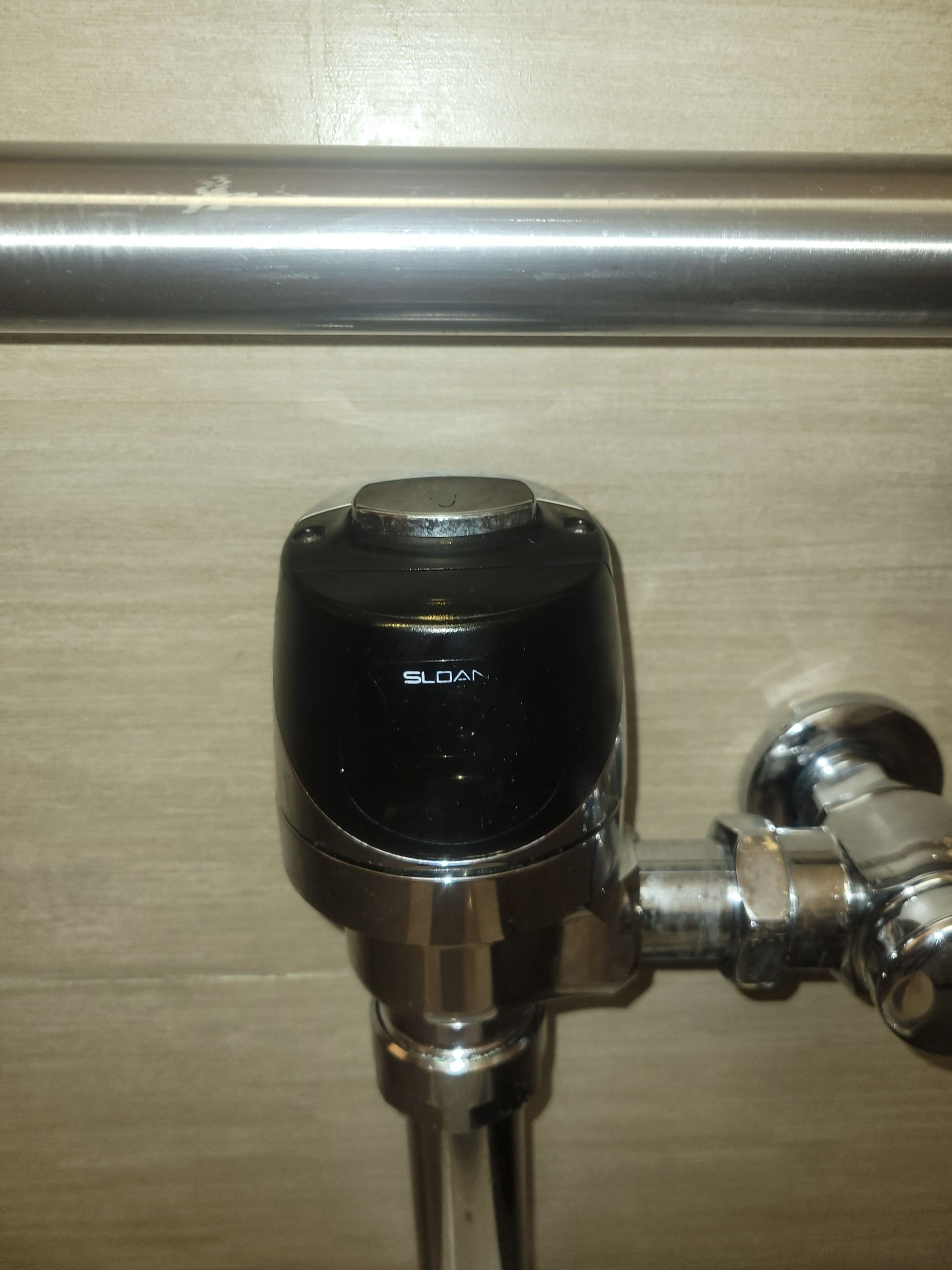 A black, cylindrical commercial toilet flush valve button mounted on a chrome pipe against a light, wood-paneled wall.