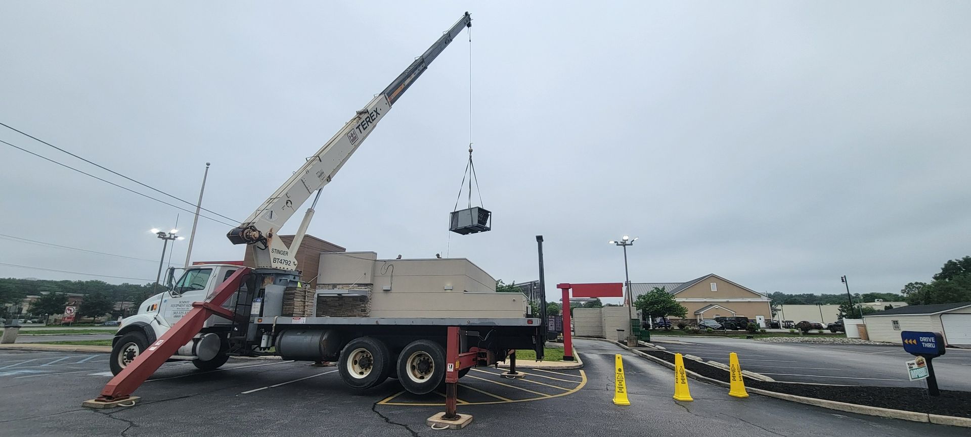 A crane truck in a parking lot lifts an HVAC unit into the air with its boom, assisted by orange safety cones.