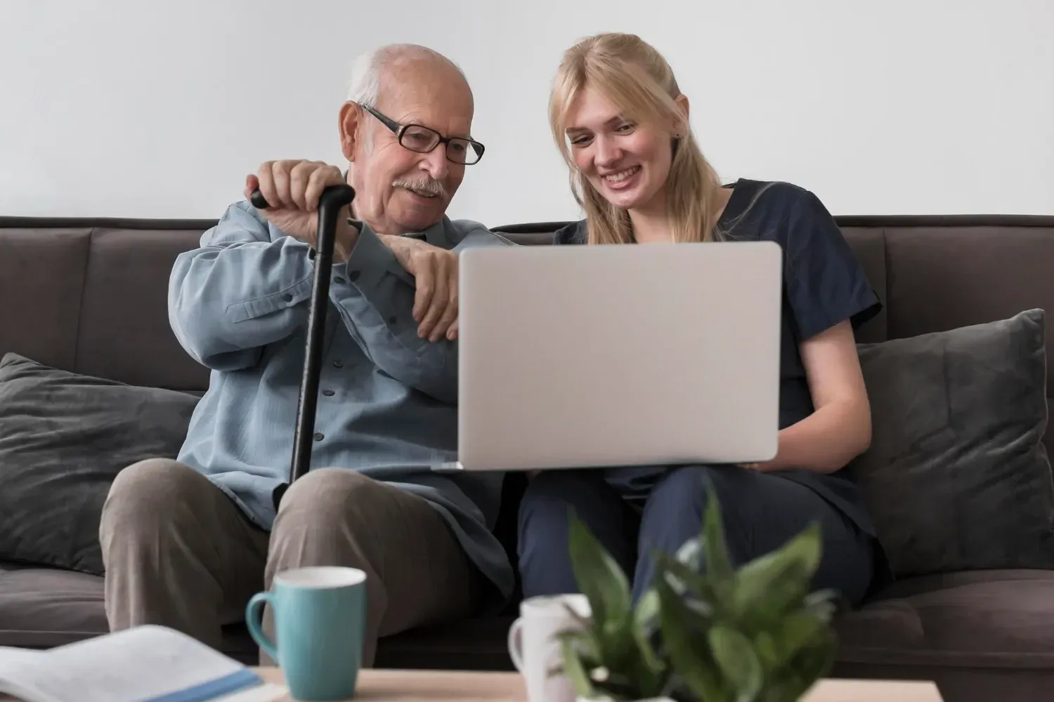 Two people smiling on a couch, looking at a laptop together, with a mug and plant in the foreground.