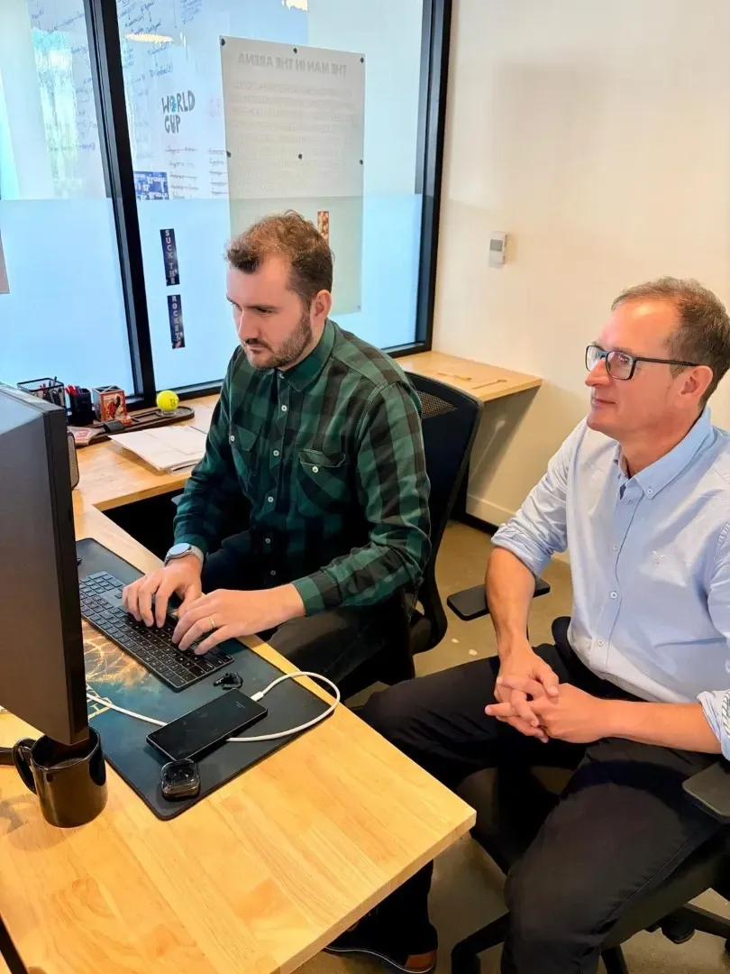 Two coworkers sitting at a desk, one typing on a laptop while the other watches in an office.