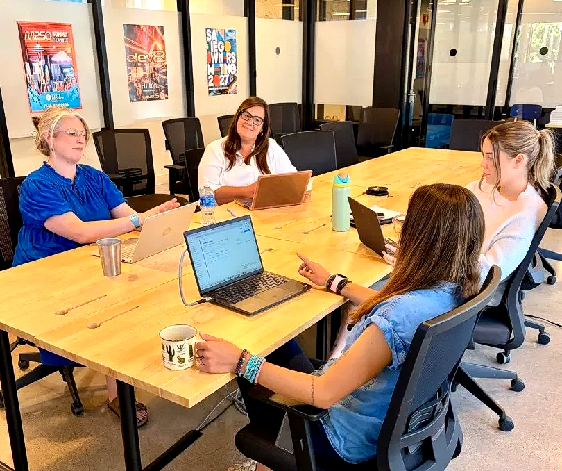 Four people in a conference room working on laptops around a wooden table.