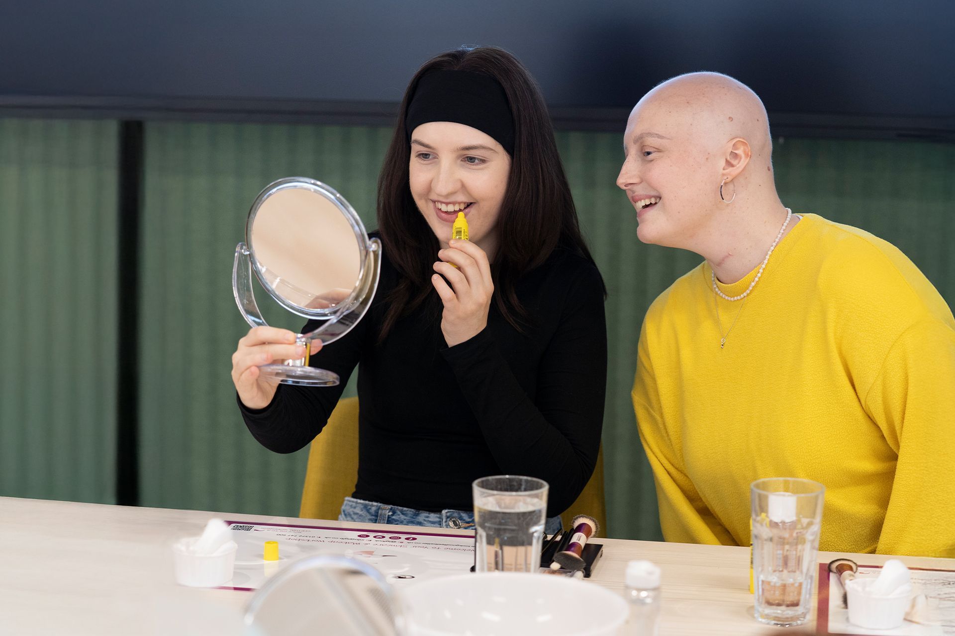 Two people at a table, one holding a mirror and lipstick, smiling in a bright room