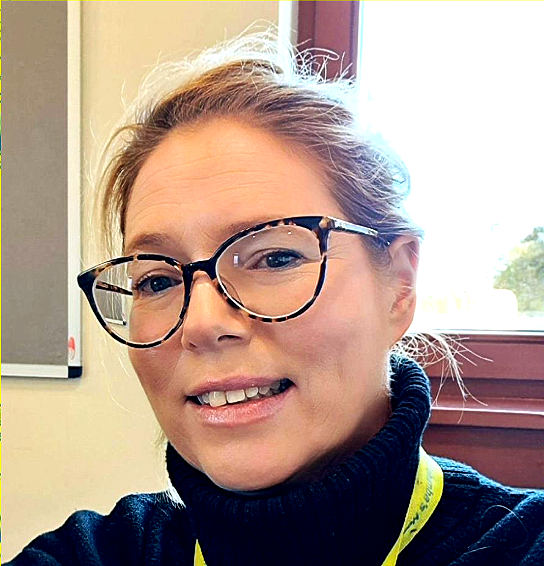 Woman wearing glasses and a black turtleneck smiles in an indoor office setting.