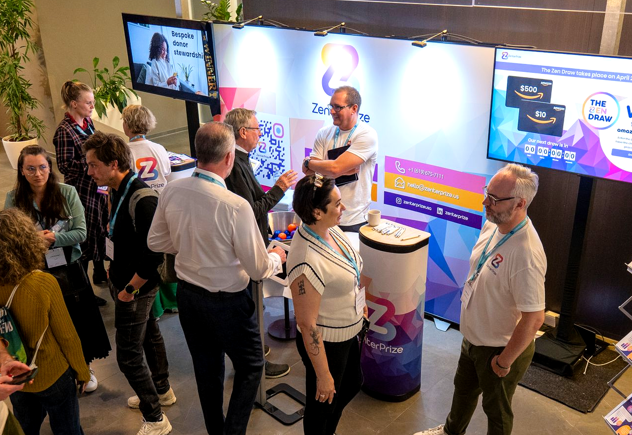 People mingling at a tech booth with two large screens in a busy conference exhibit area