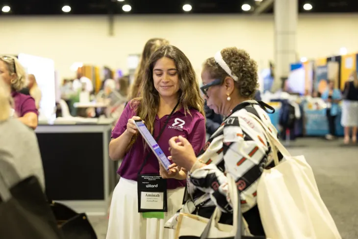 Two women at a conference booth, one showing a purple item to the other in a busy event hall.