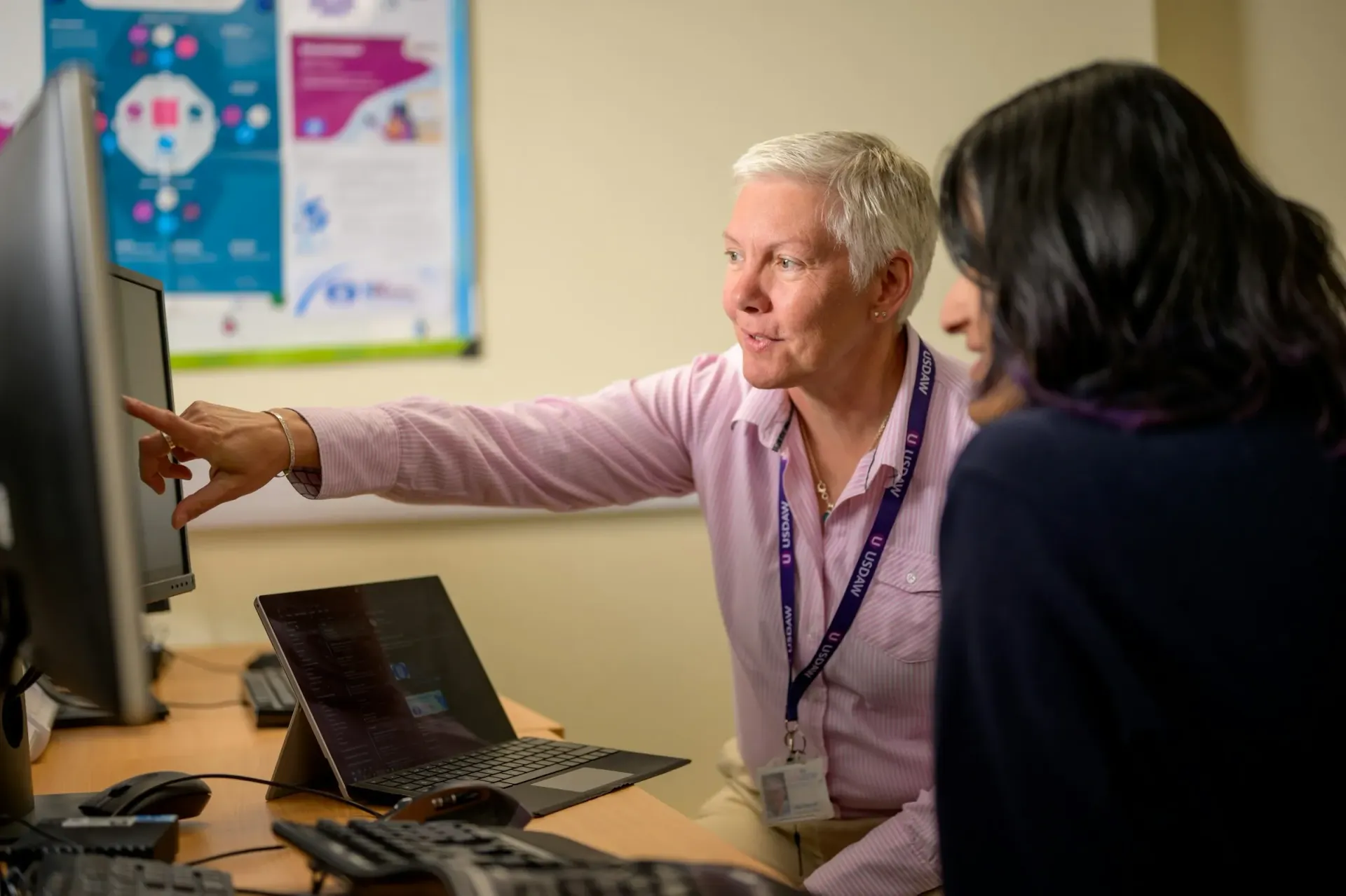 Woman in a lavender shirt points at a desktop monitor during a discussion in an office.