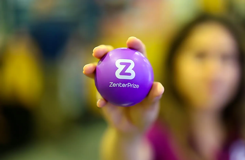 Hand holding a purple Zenoti-branded ball in front of a blurred background and person
