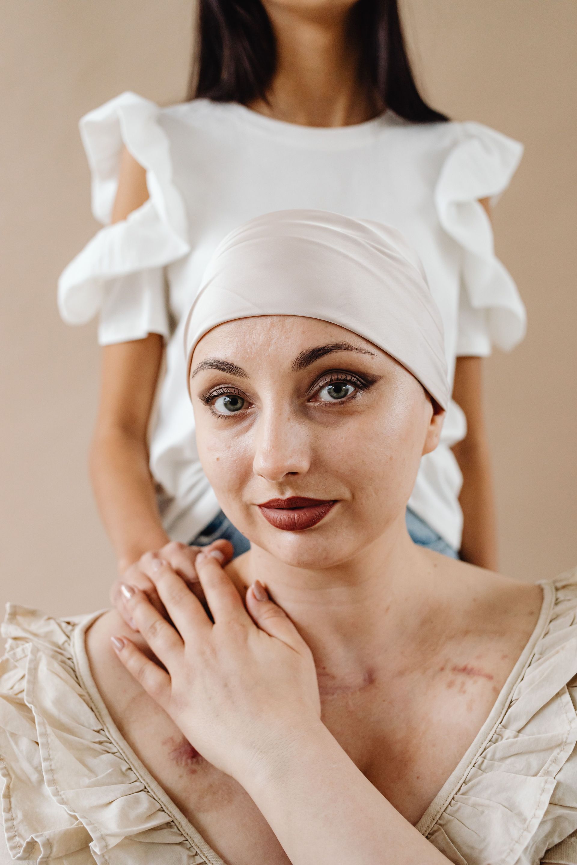 Woman with wrapped head and chest scars, being comforted by another person in a white dress