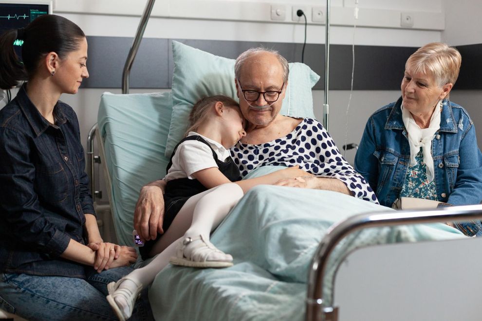 Three visitors sit beside a child resting in a hospital bed, with an older patient smiling nearby.