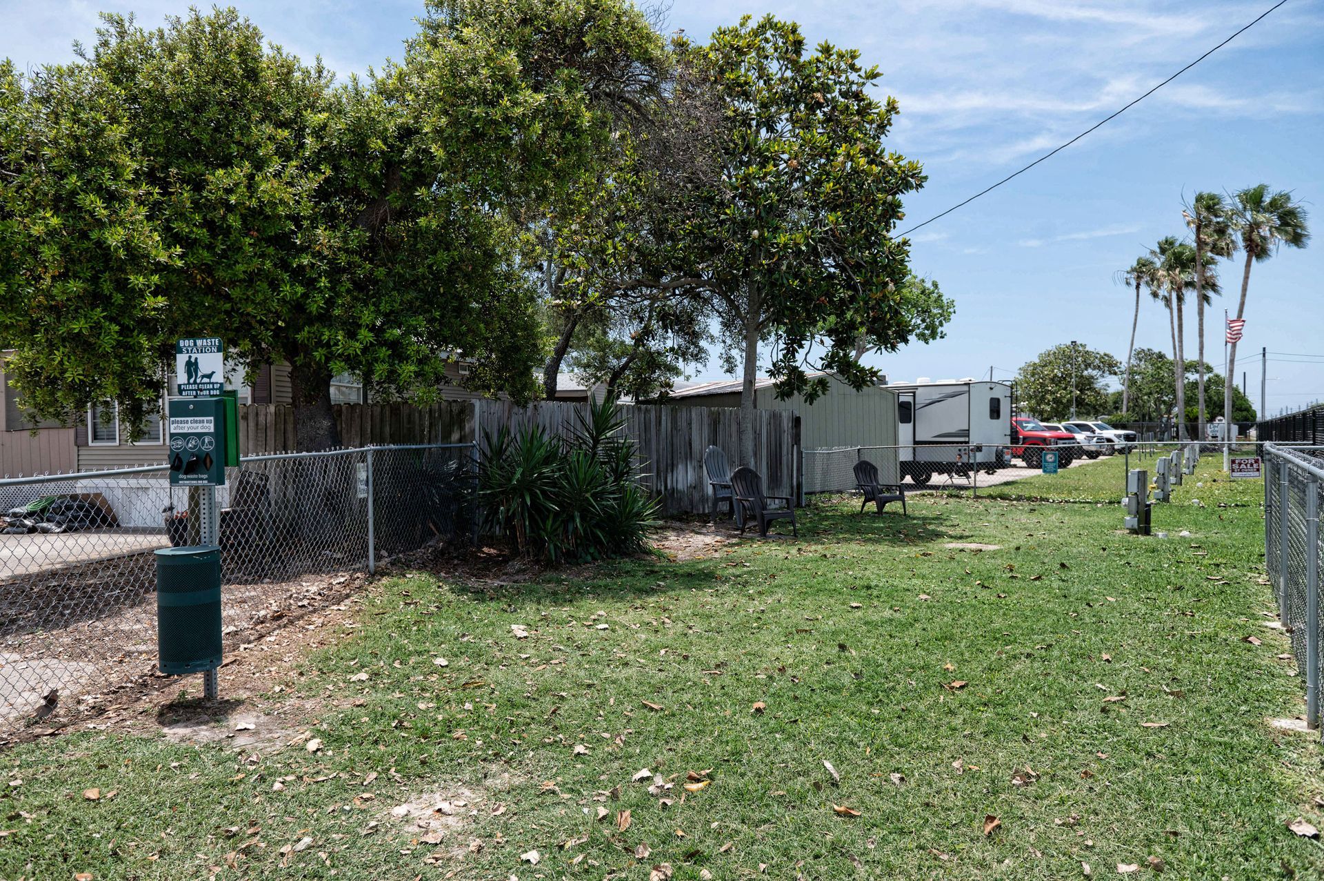 Grassy roadside lot with a chain-link fence, trees, and parked vehicles under a sunny sky
