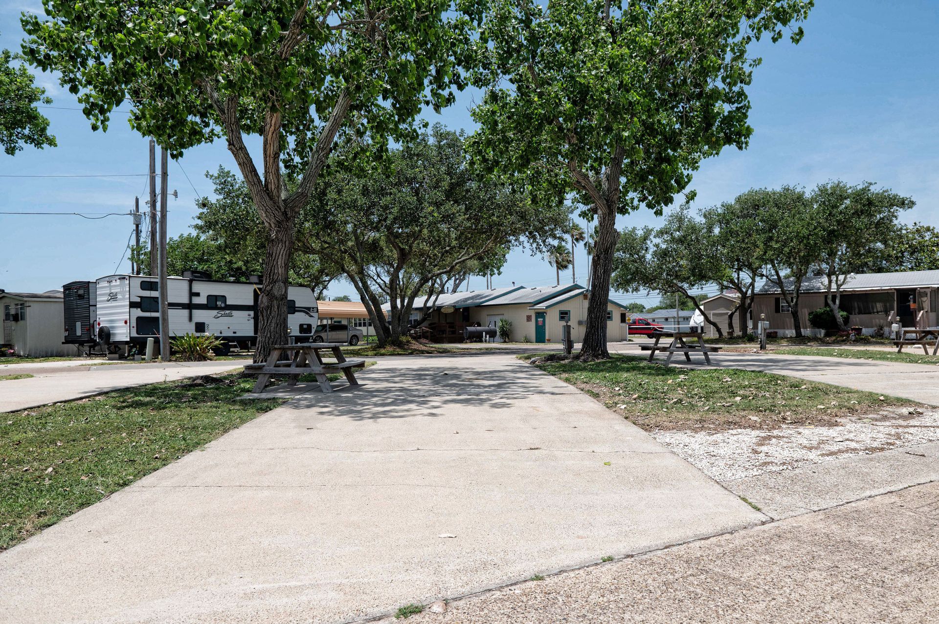 Paved walkway through a park with large trees, picnic tables, and houses in the background