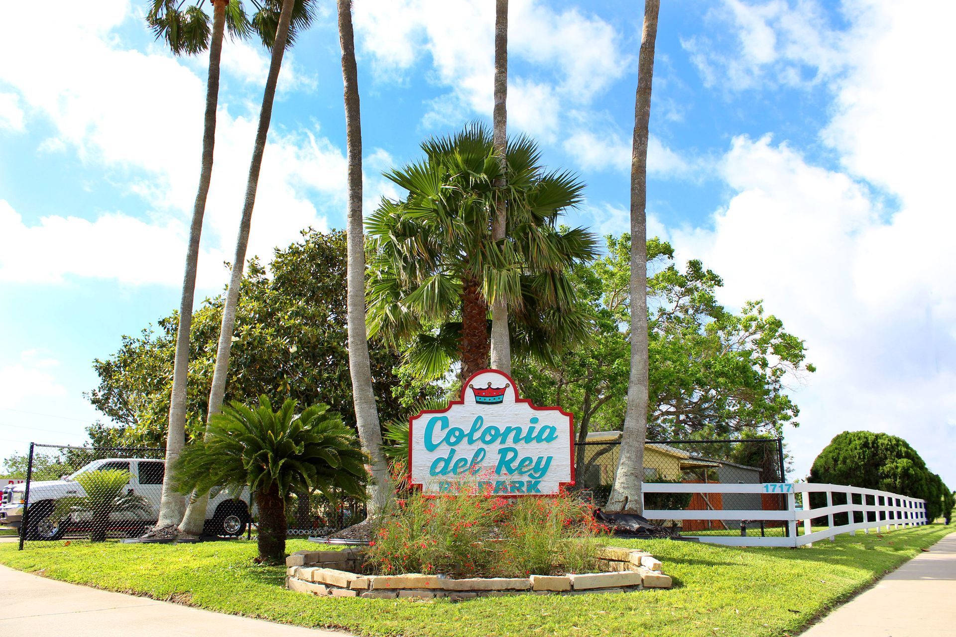 Entrance sign for Colonia del Rey with palm trees, green landscaping, and a white fence under a blue sky
