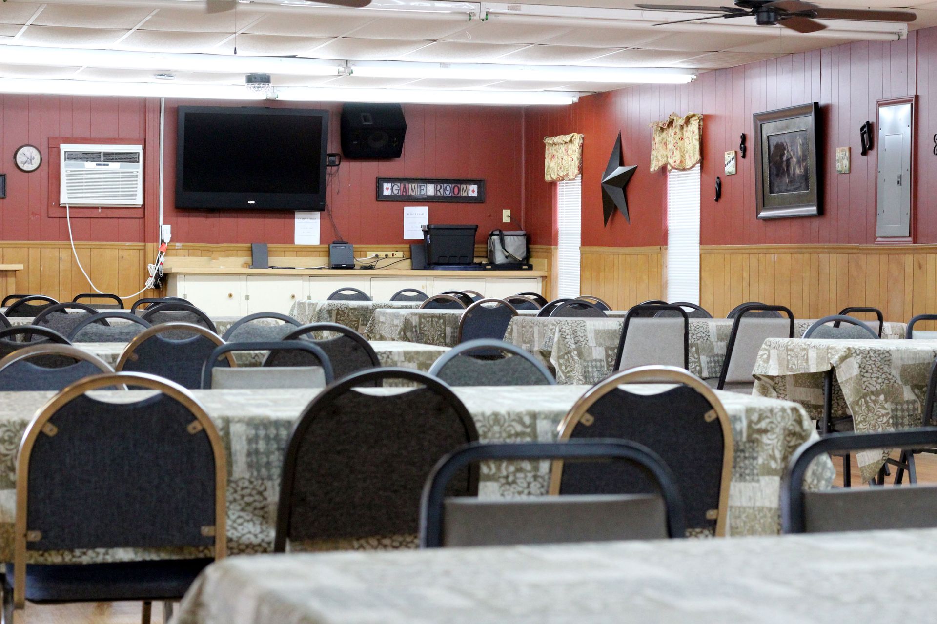 Empty banquet hall with rows of chairs and tables, TV and wood-paneled stage area at the front.